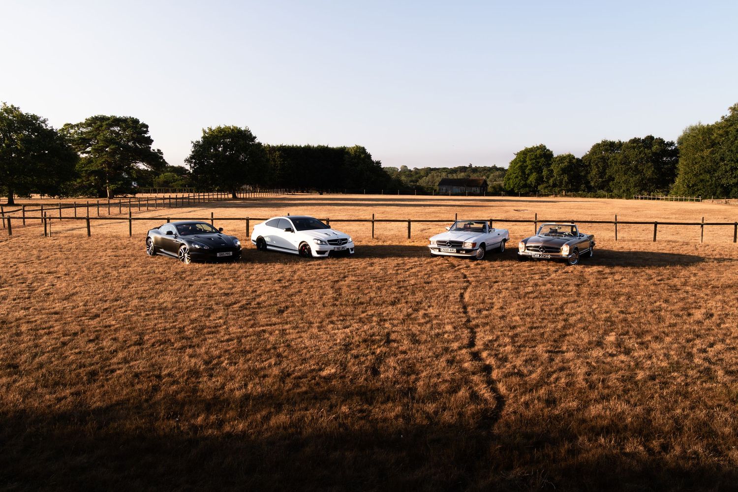 Four luxury sports cars parked in a golden field at sunset with trees and wooden fencing in the background.