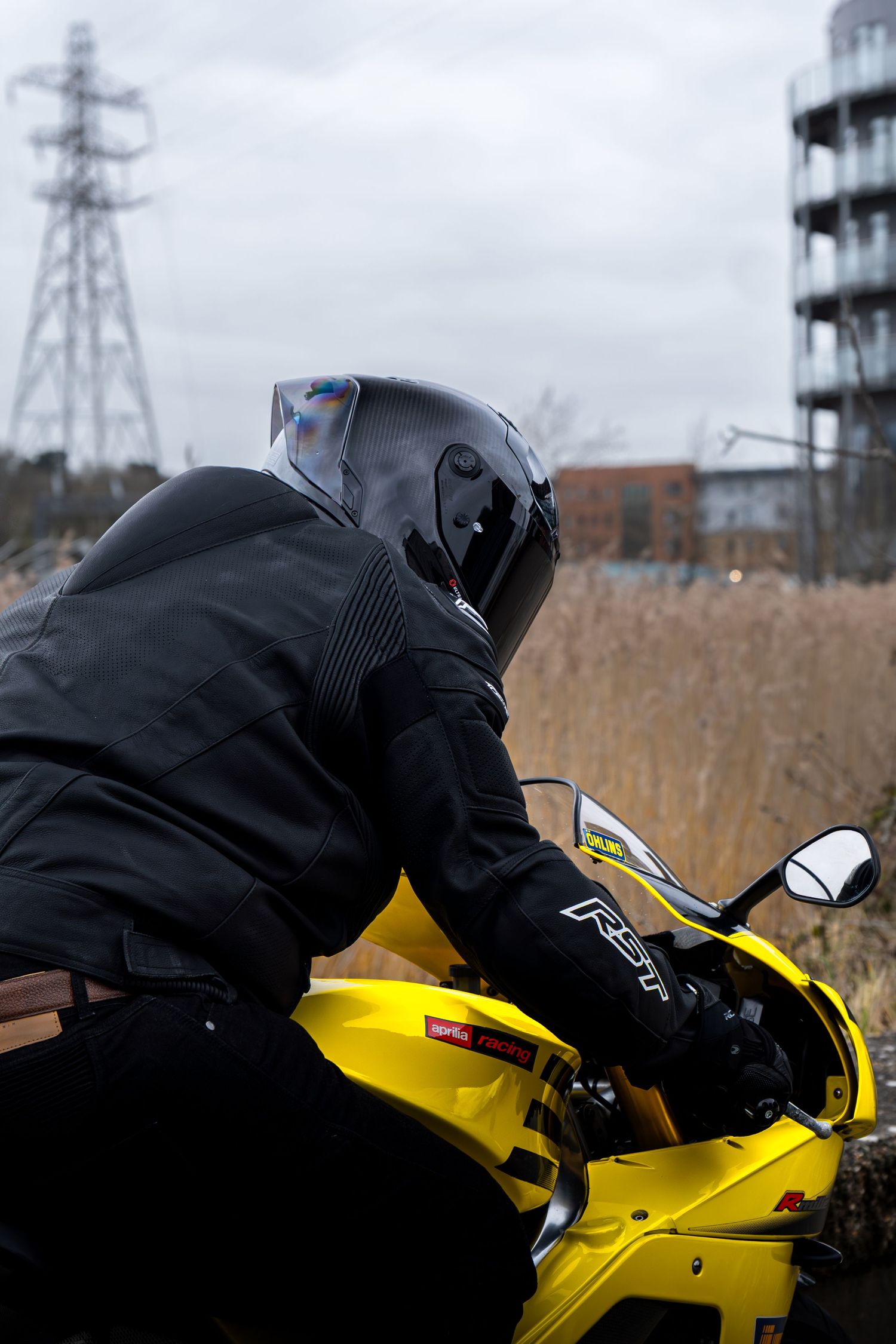 Rear view of a motorcyclist in black RST gear seated on a yellow Aprilia near tall reeds.