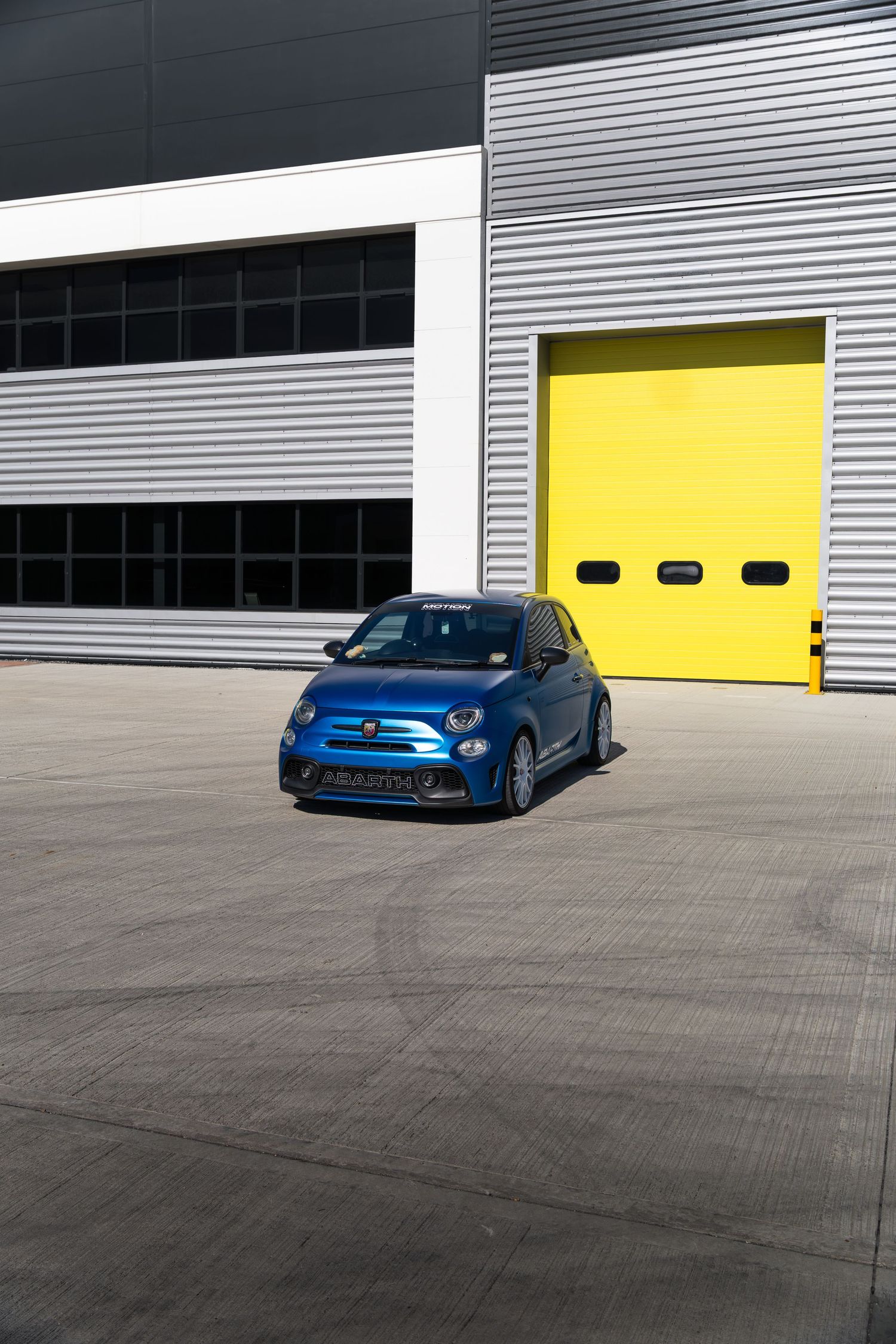 Blue Abarth 595 parked outside industrial building with bright yellow roller shutter door.