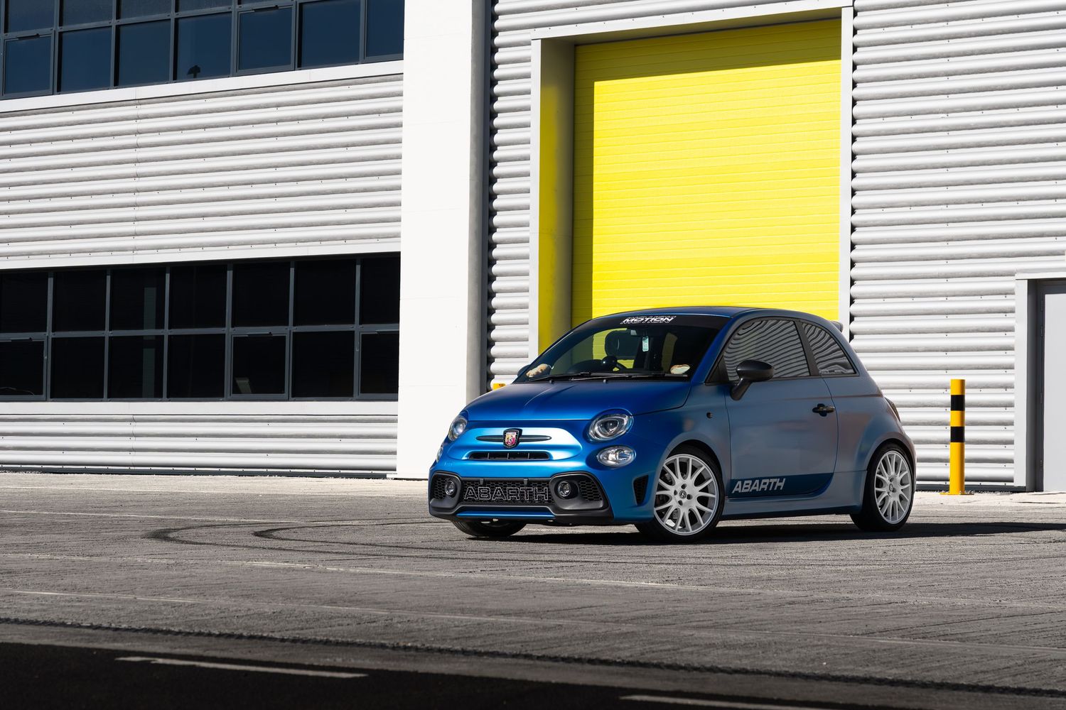 Blue Abarth 595 tuned hatchback parked beside yellow garage door at motorsport facility.