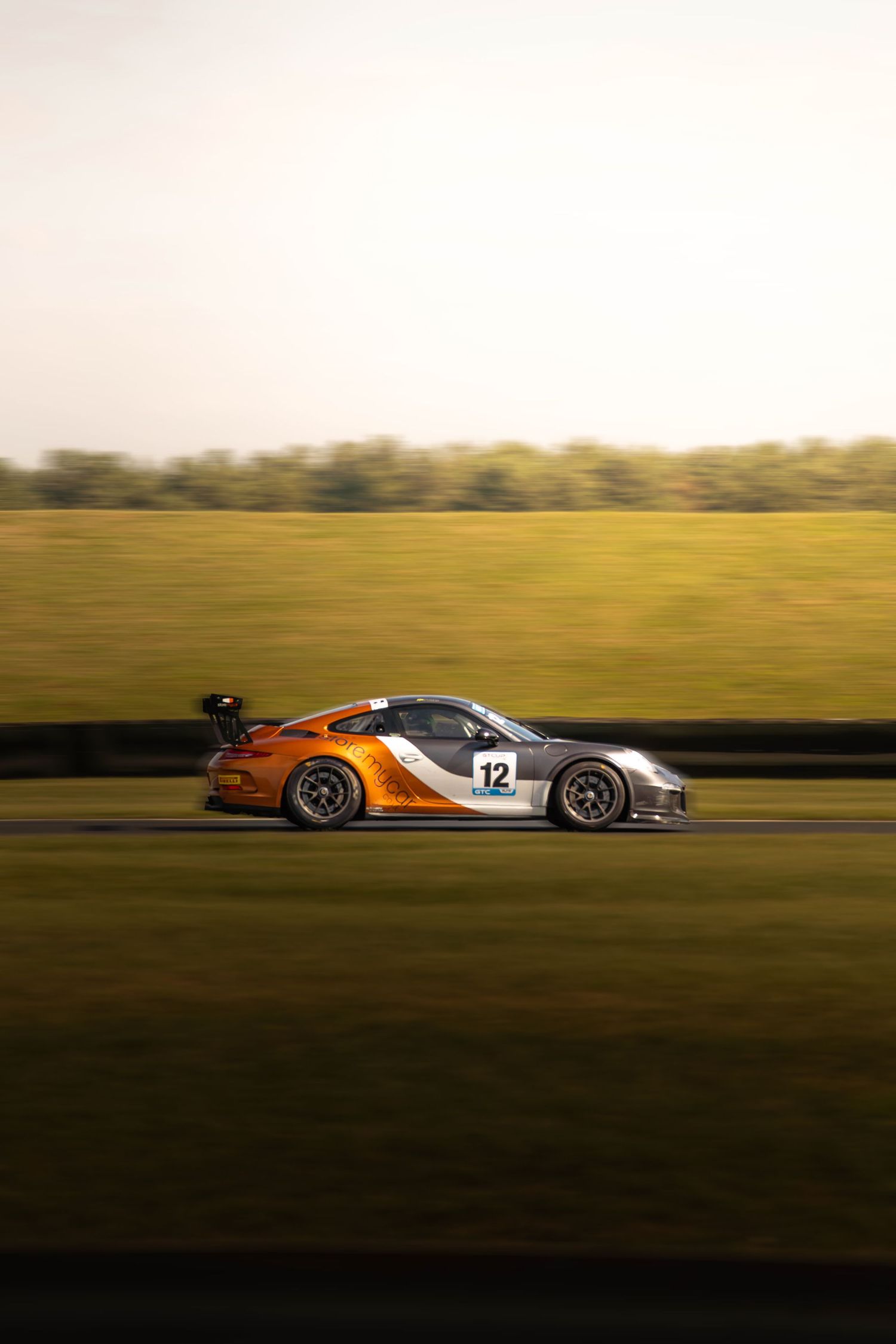 Orange and silver Porsche 911 GT3 number 12 racing on a motorsport circuit.