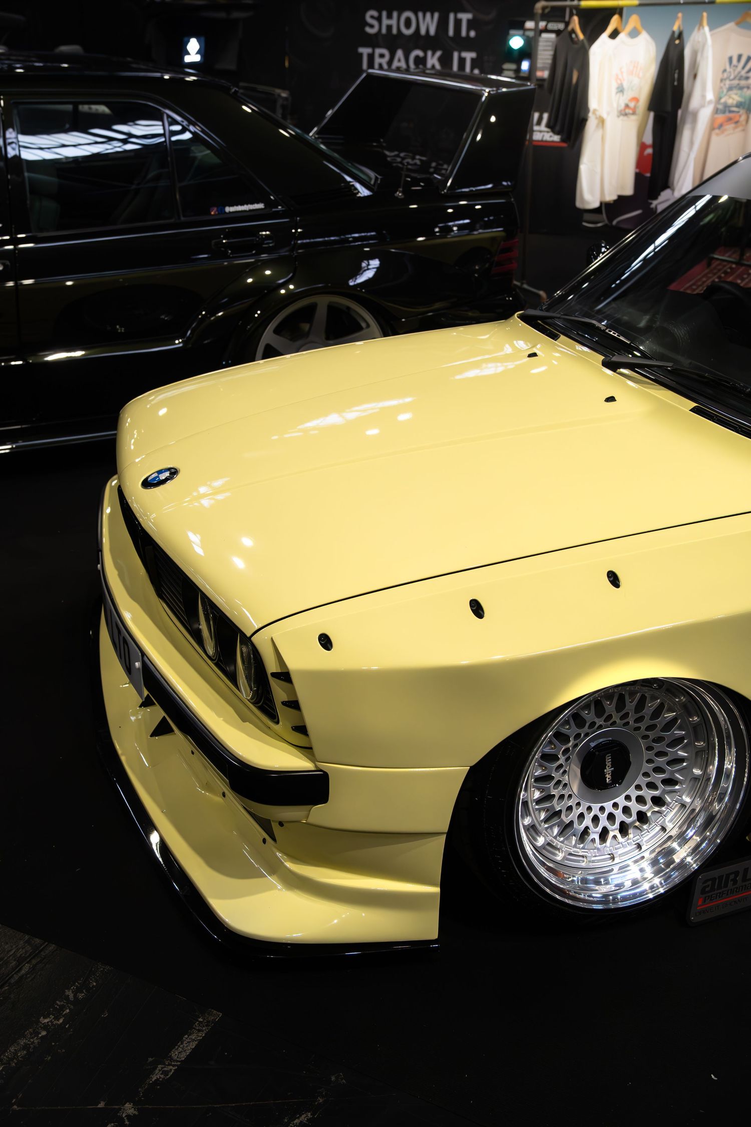 Pale yellow widebody BMW E30 with BBS wheels on display at an indoor automotive exhibition.