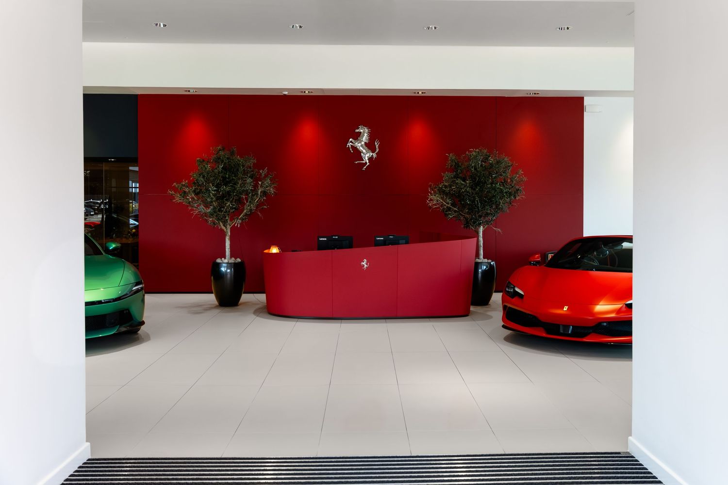 Ferrari dealership showroom with red reception desk, prancing horse logo, two sports cars, and decorative olive trees.