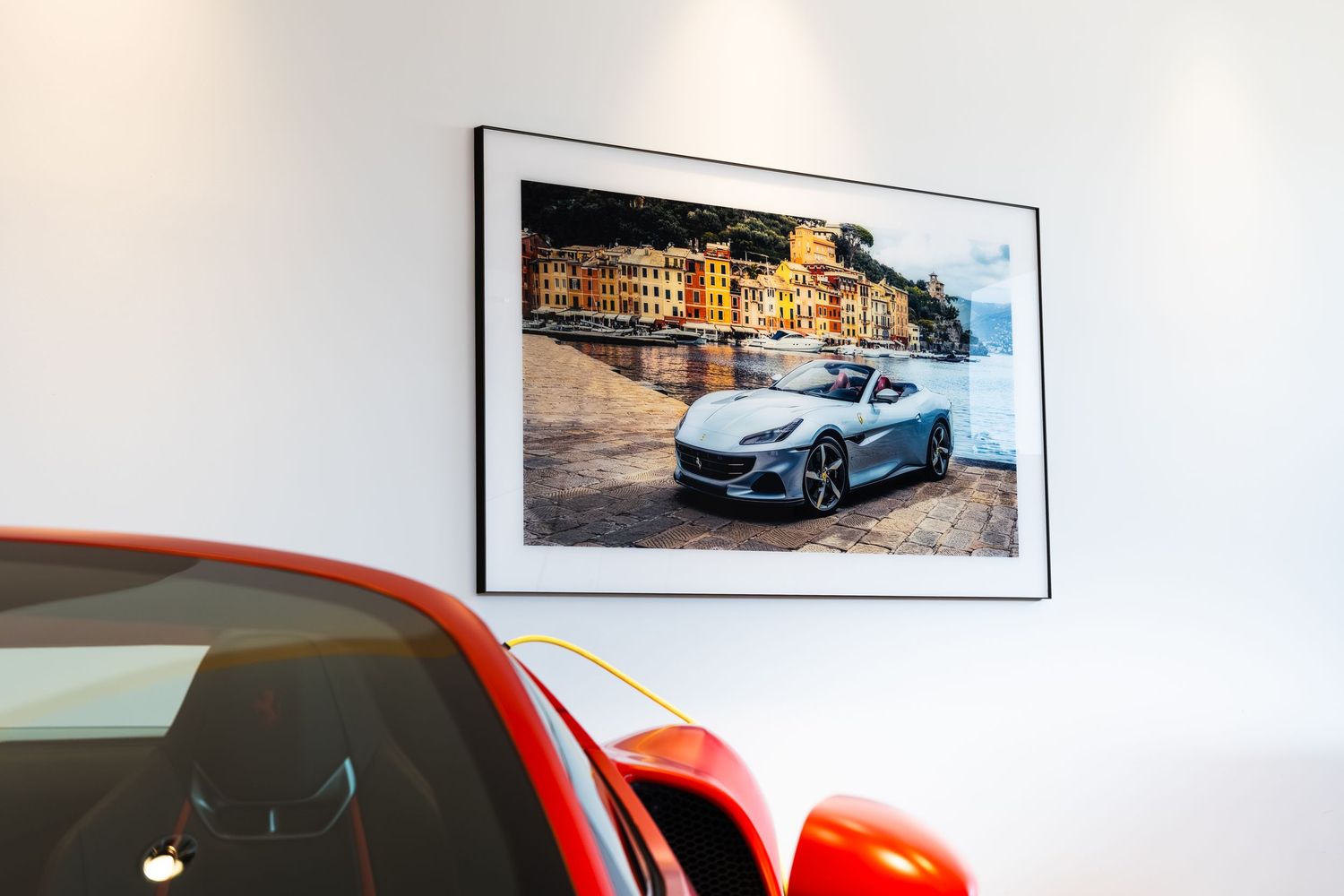 Red sports car displayed in showroom beside framed photo of silver Maserati GranTurismo in Portofino, Italy.