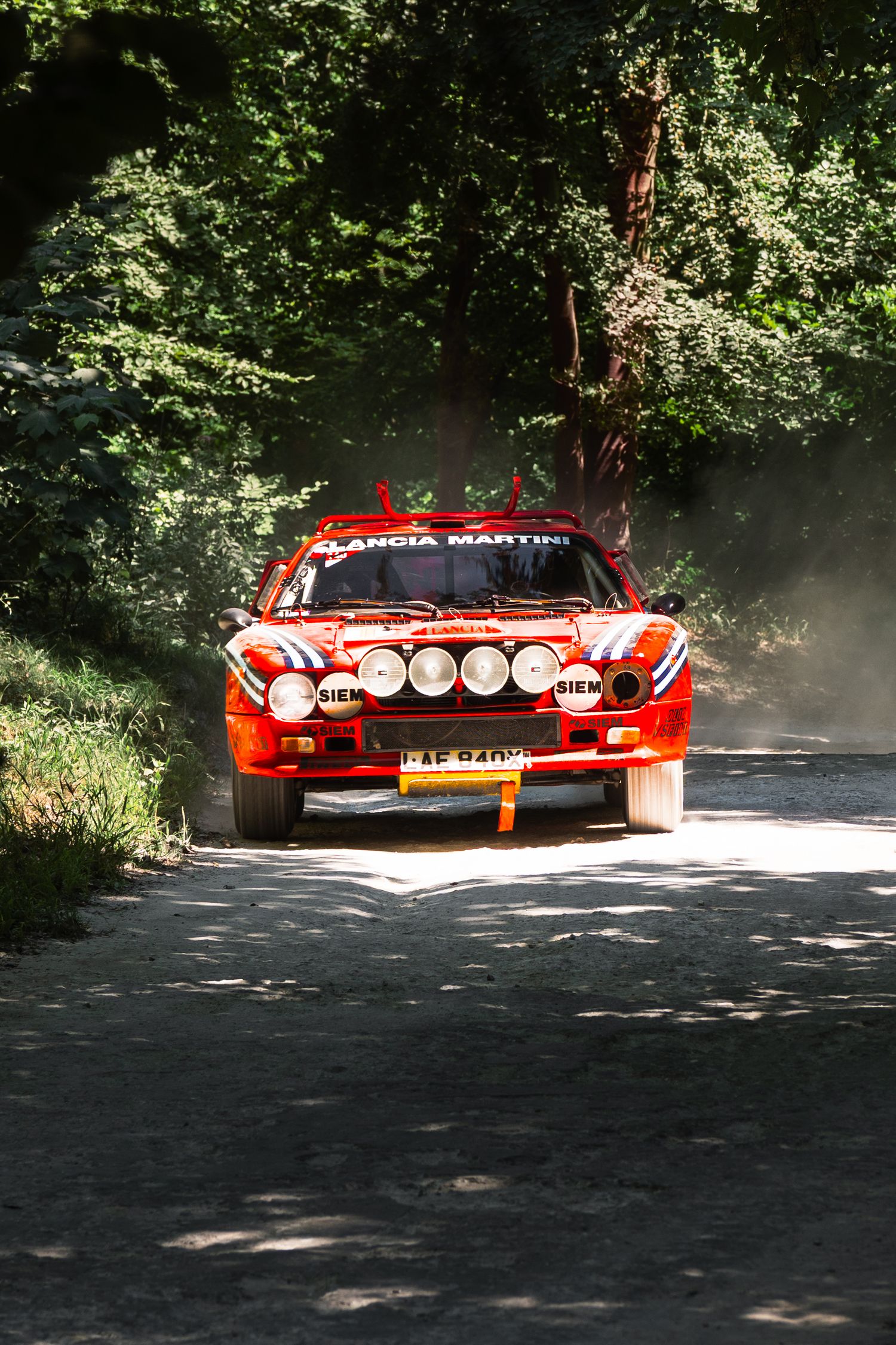 Red Lancia Martini rally car racing on a gravel forest stage road with dust trailing behind.