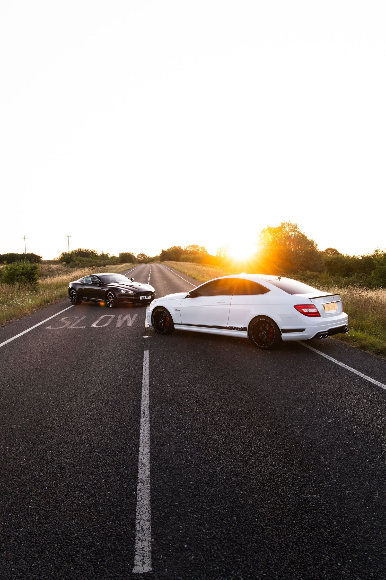 A white Mercedes coupe and black sports car parked on a rural road at golden sunset with SLOW marking visible.