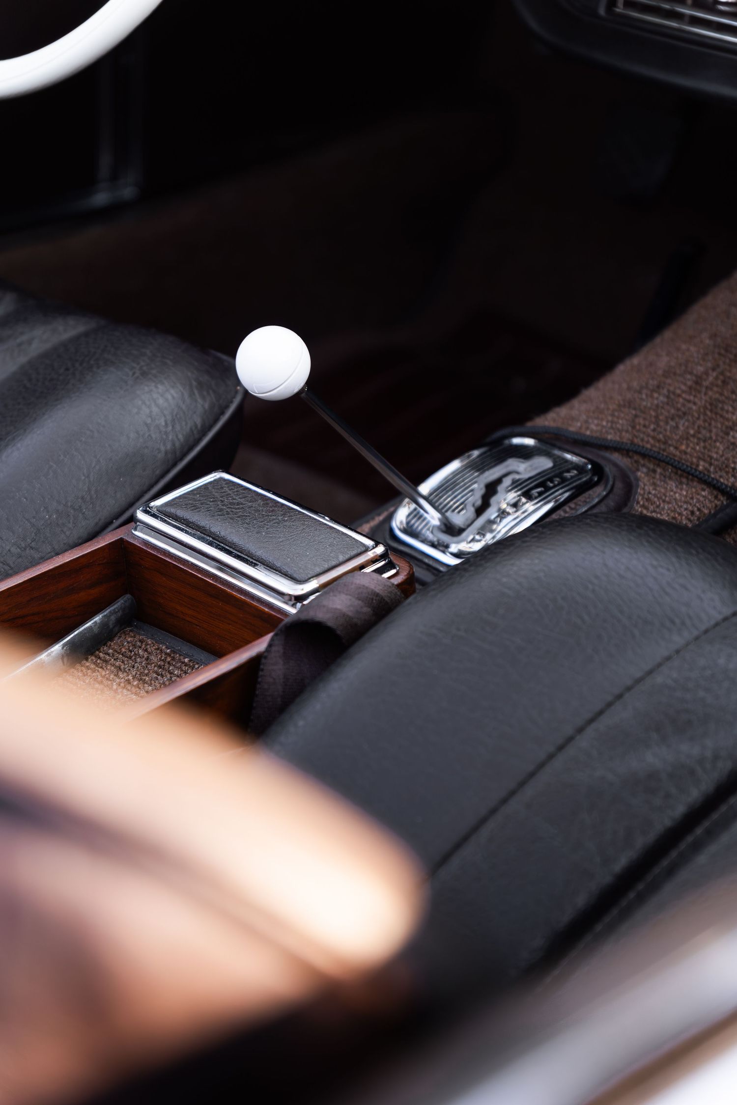 Close-up of a classic car interior featuring a white ball gear shift, wooden trim, and black leather seats.
