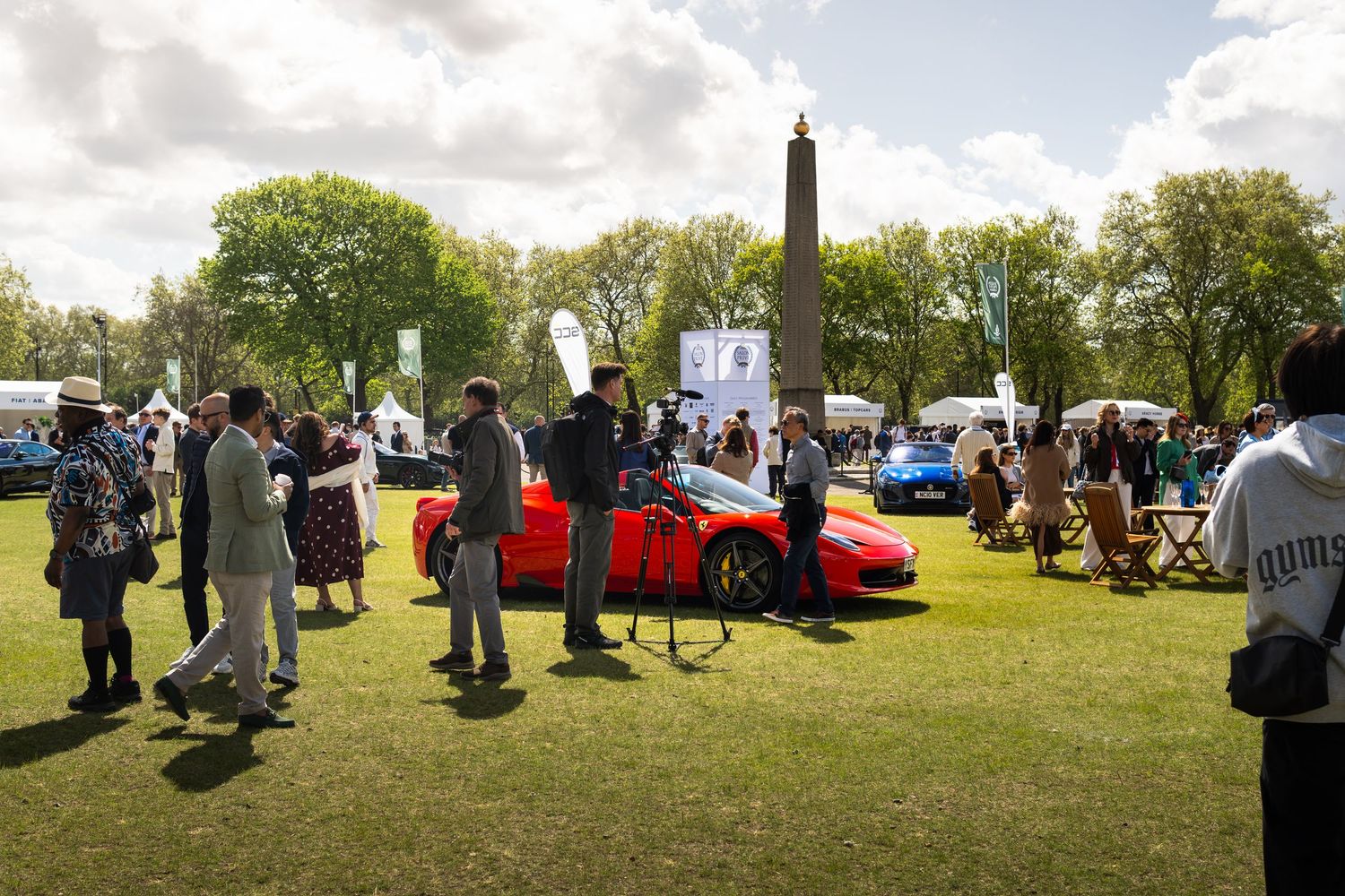 Red Ferrari sports car surrounded by elegantly dressed spectators at an outdoor luxury car show.