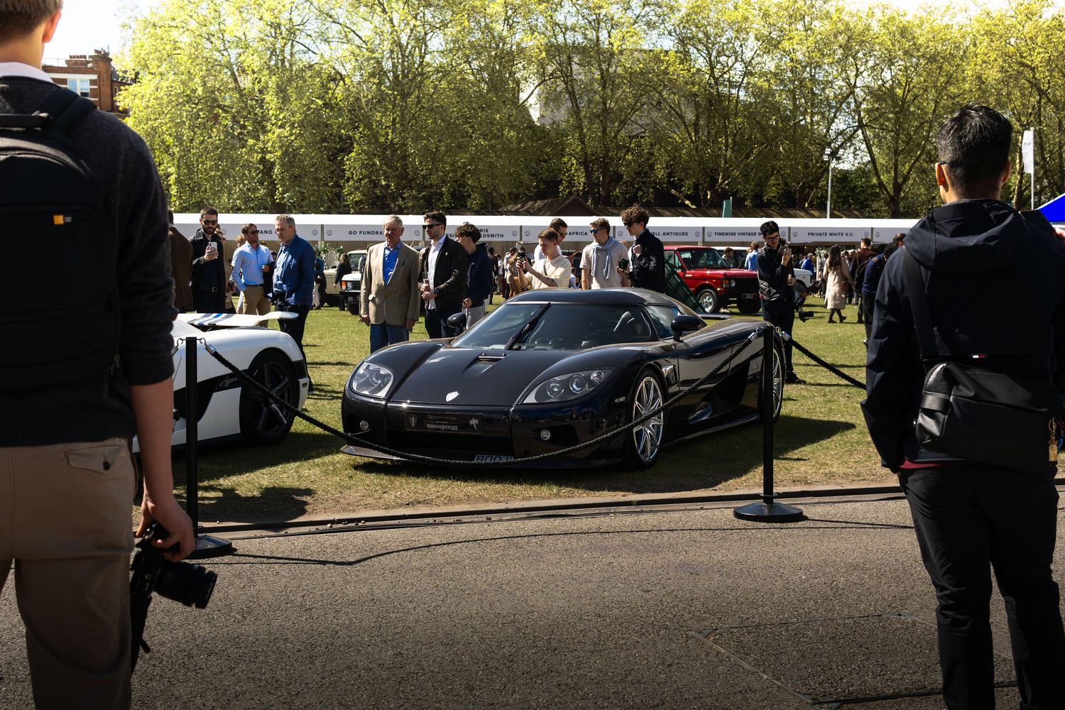 Dark carbon fiber Koenigsegg supercar on display at an outdoor exotic car show with spectators.