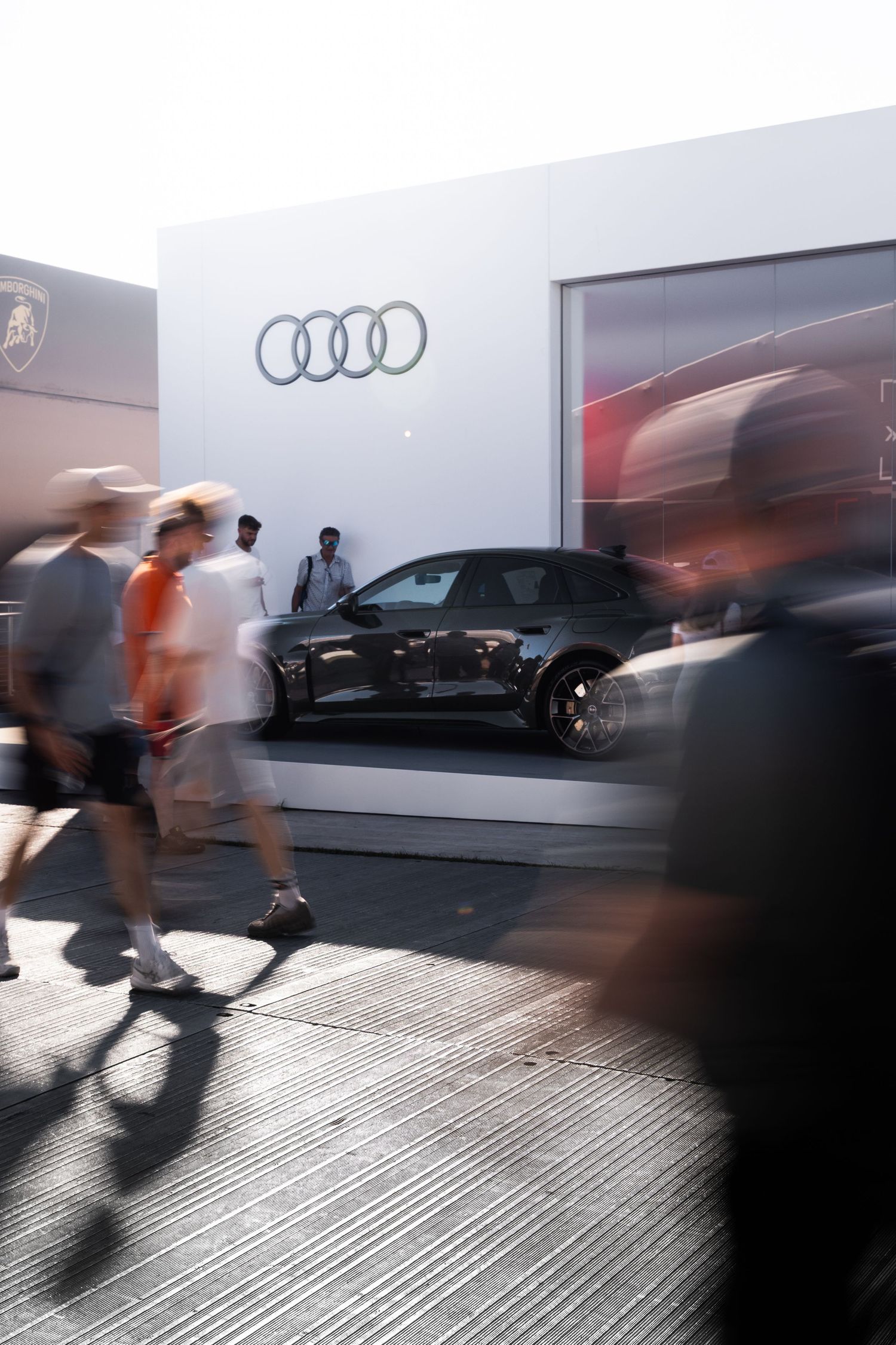 Blurred pedestrians walk past an Audi car display booth featuring a sleek black vehicle at an outdoor auto show.
