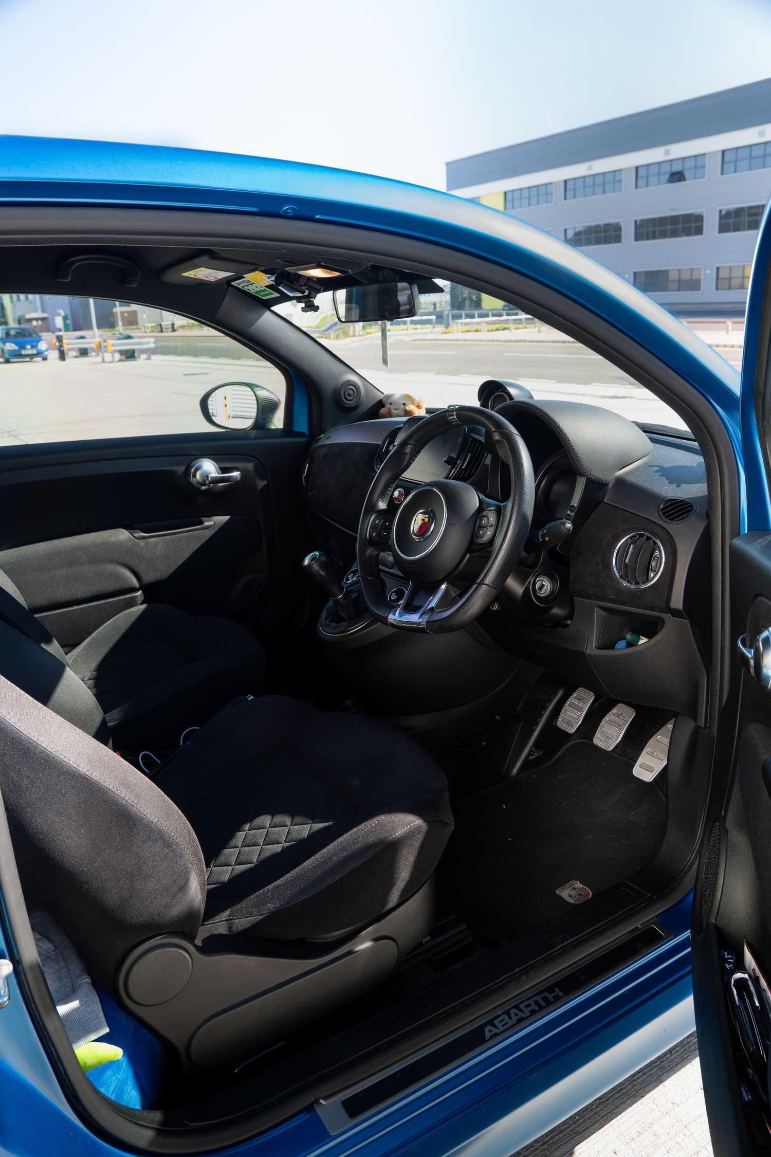 Open door view of a blue Abarth 595 revealing the black sport interior, pedals, and Abarth sill plate.