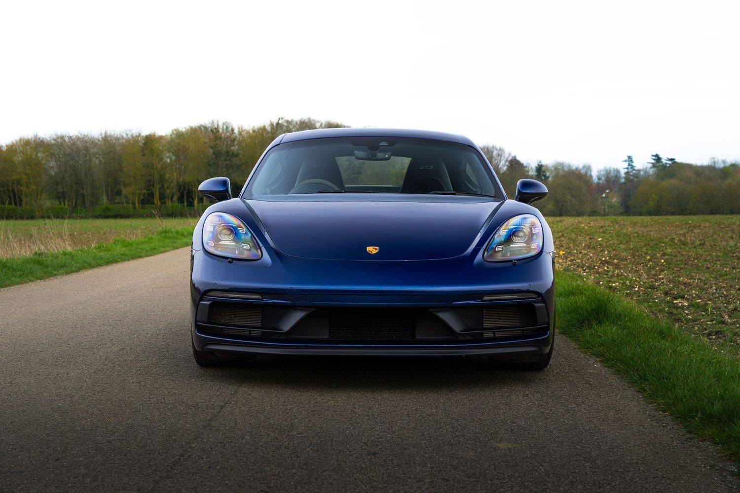Front view of blue Porsche 718 Cayman on a straight country road flanked by green trees.