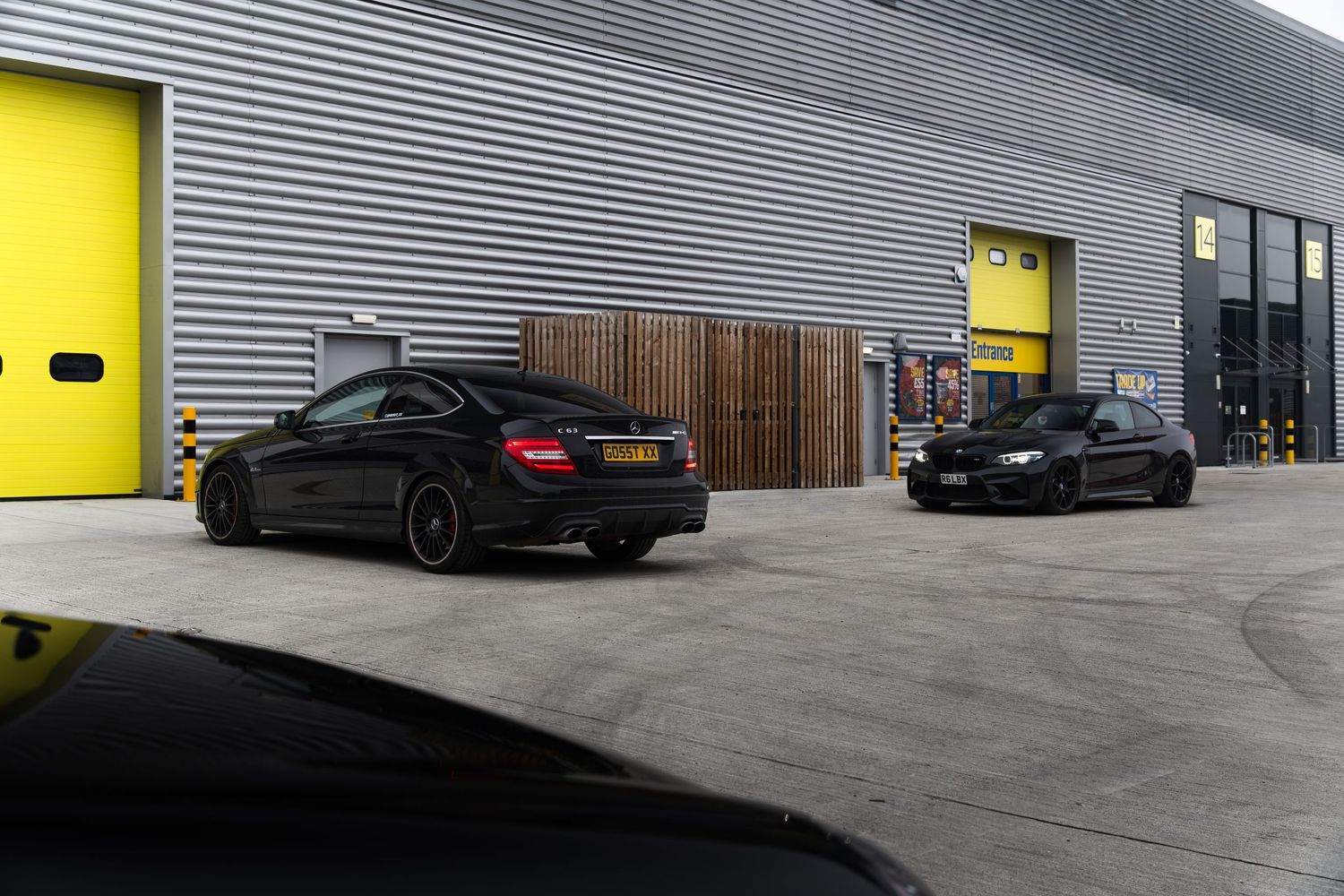 Black Mercedes-Benz C63 AMG and BMW M2 parked outside an industrial unit with yellow doors.