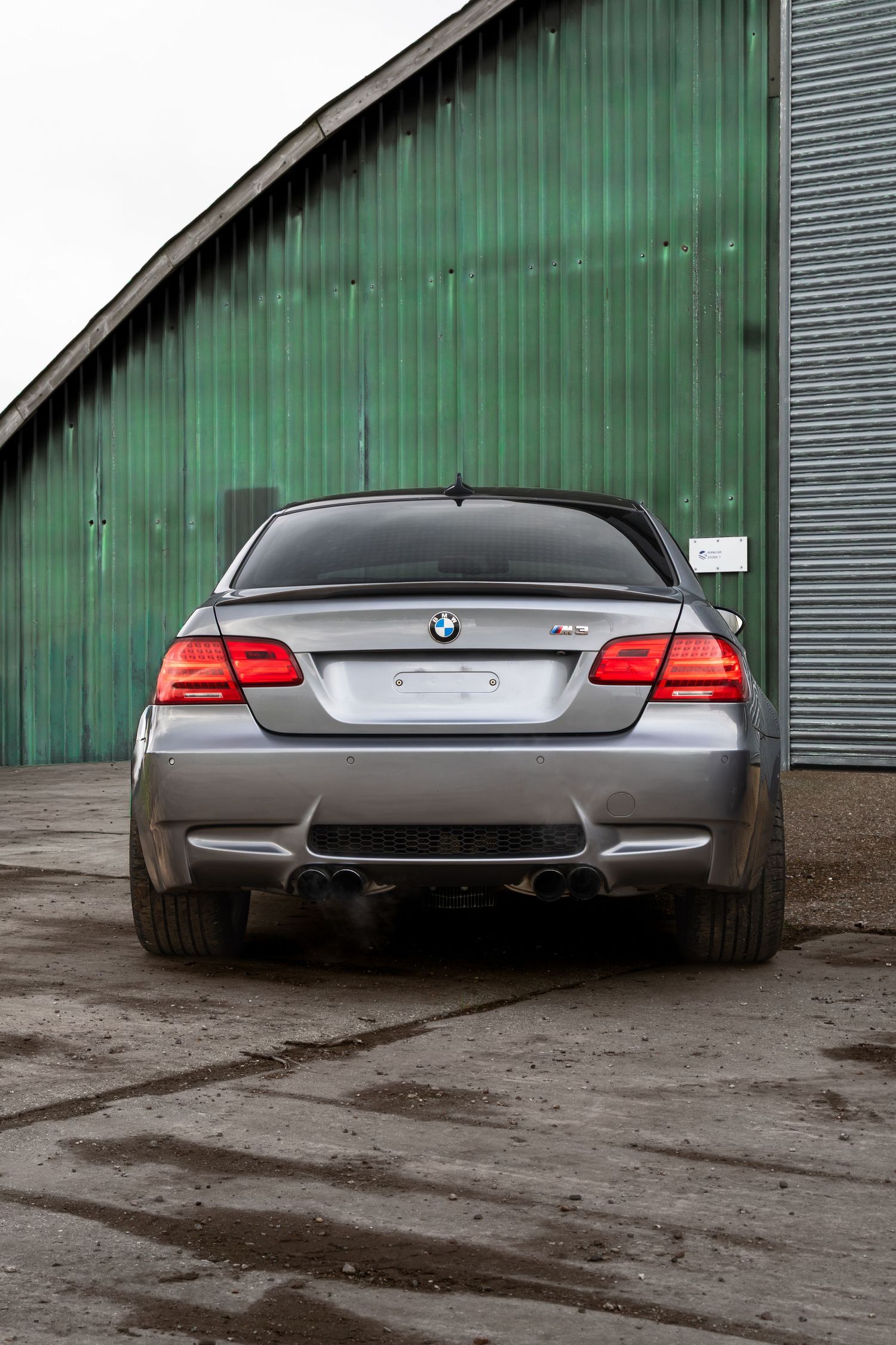 Rear close-up of a silver BMW M3 coupe parked against a green corrugated metal building.
