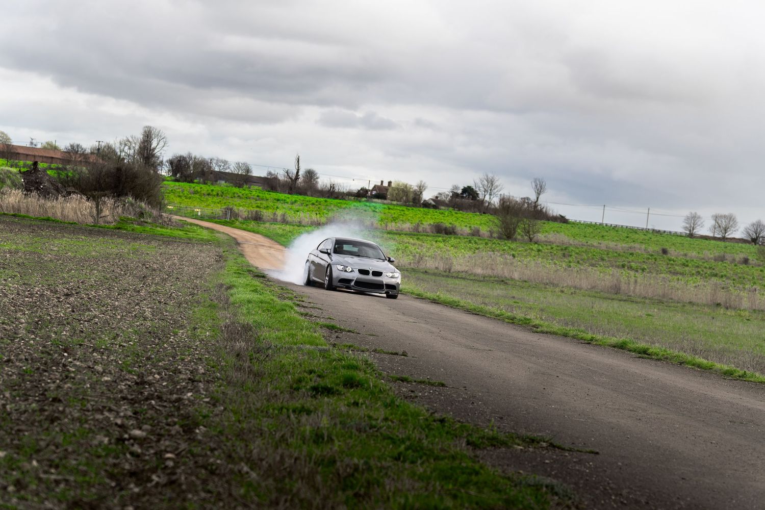 Silver BMW M3 drifting on a rural road leaving tire smoke trails on an overcast day.