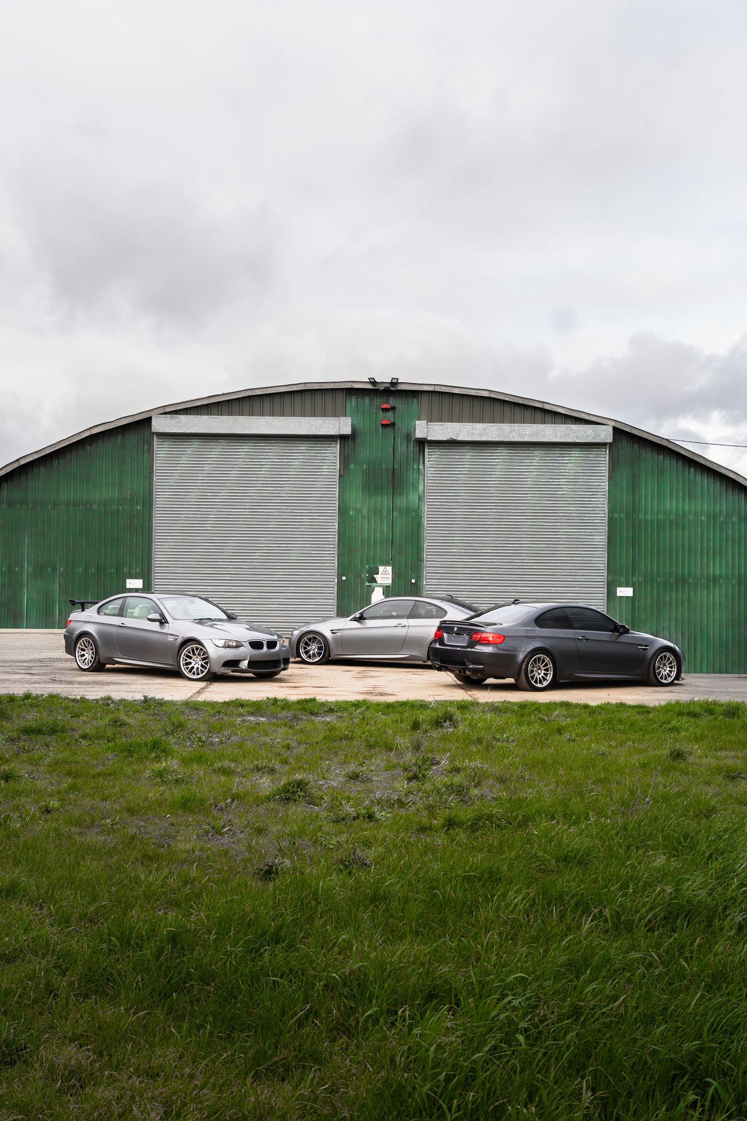 Rear view of three grey BMW M3 coupes lined up outside a green corrugated metal hangar.
