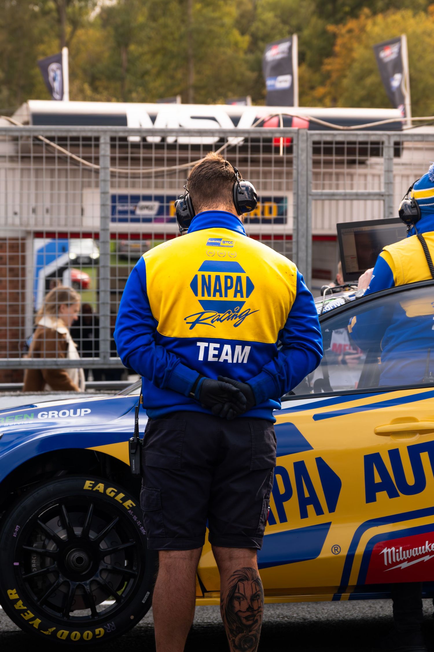 NAPA Racing Team mechanic in blue and yellow jacket standing beside race car at BTCC event.