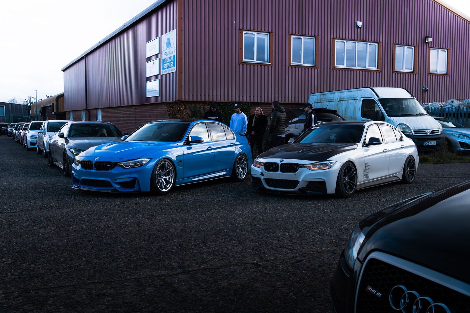 Row of modified BMW and Audi cars parked outside an industrial warehouse building.