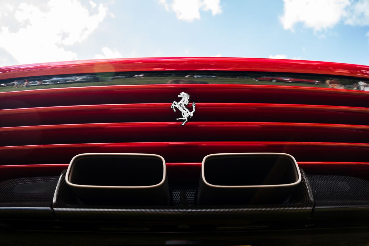 Close-up of a red Ferrari rear grille with prancing horse logo and dual exhaust pipes against a cloudy sky.