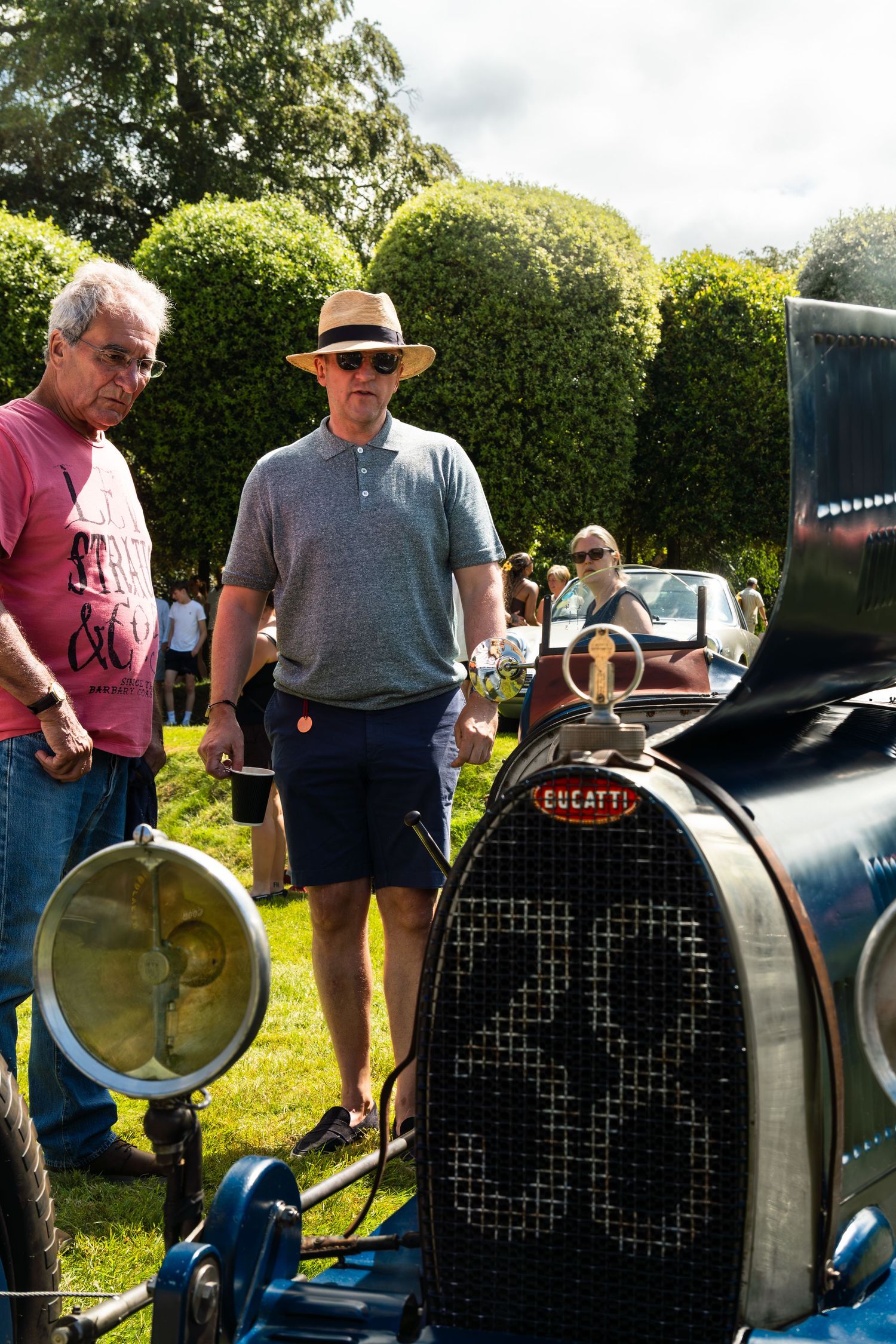 Two men admiring a vintage Bugatti car at an outdoor classic car show on a sunny day.