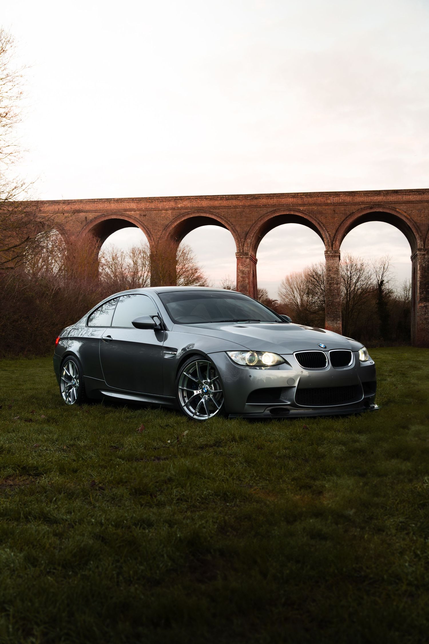Silver BMW M3 coupe parked on grass before a brick viaduct at dusk.