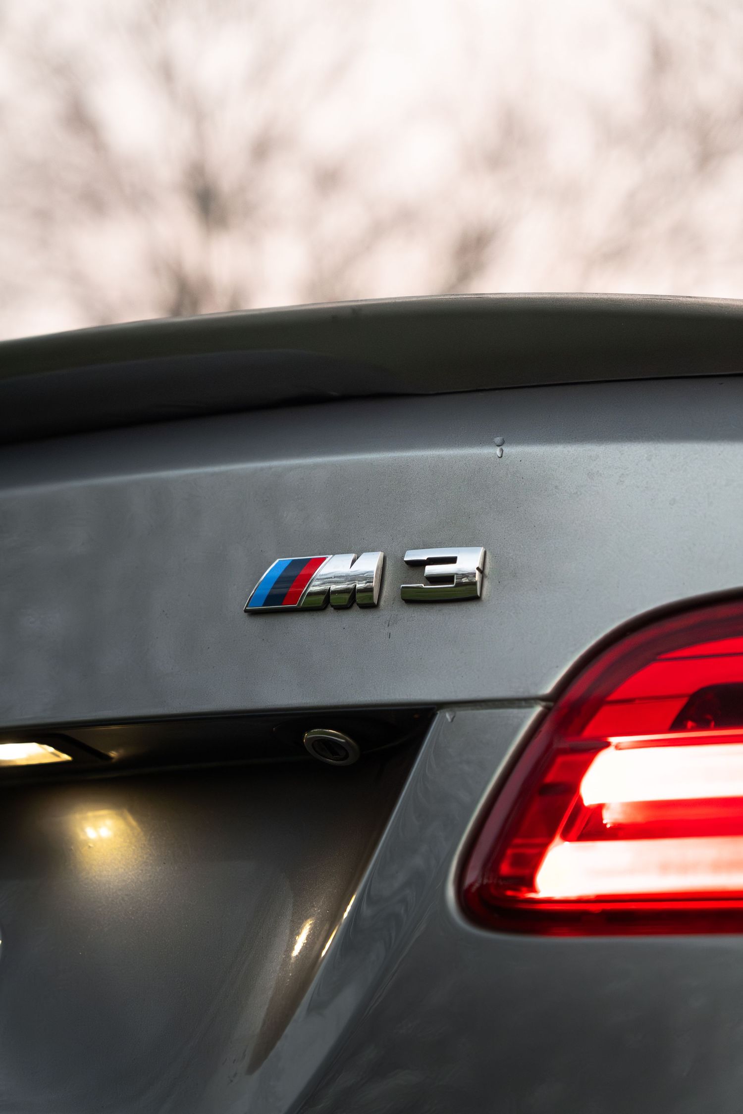 BMW M3 badge on the trunk lid of a grey coupe with red tail light visible.