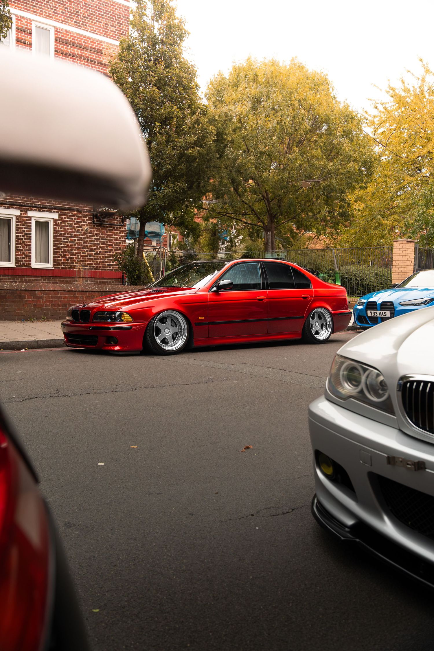 A slammed red BMW E39 5 Series parked on a residential street among other modified cars.