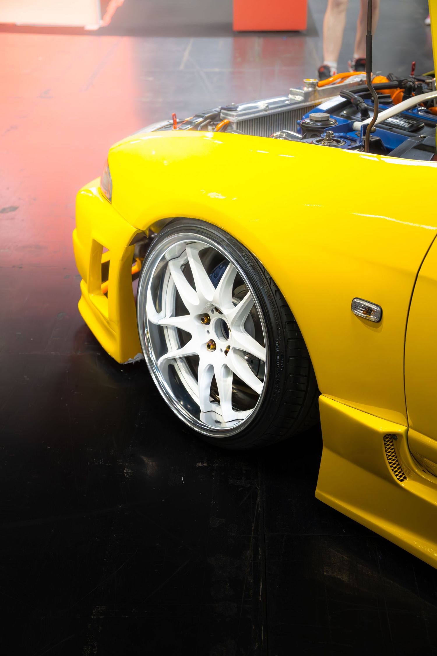 Yellow modified Japanese sports car with white alloy wheels and open hood at car show.