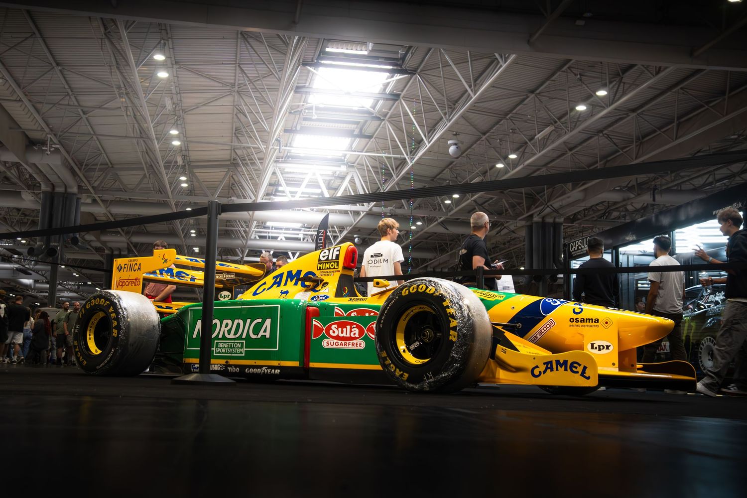 Yellow and green Benetton Camel Formula 1 car on display at an indoor motorsport show.