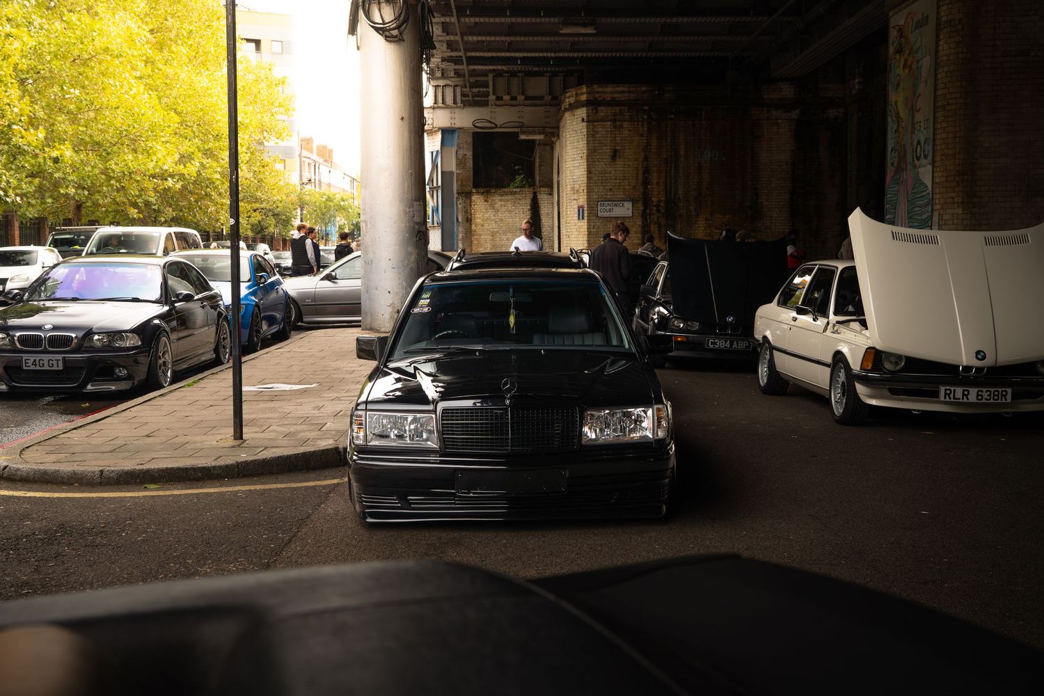 Black Mercedes-Benz W124 parked under urban overpass surrounded by classic cars.