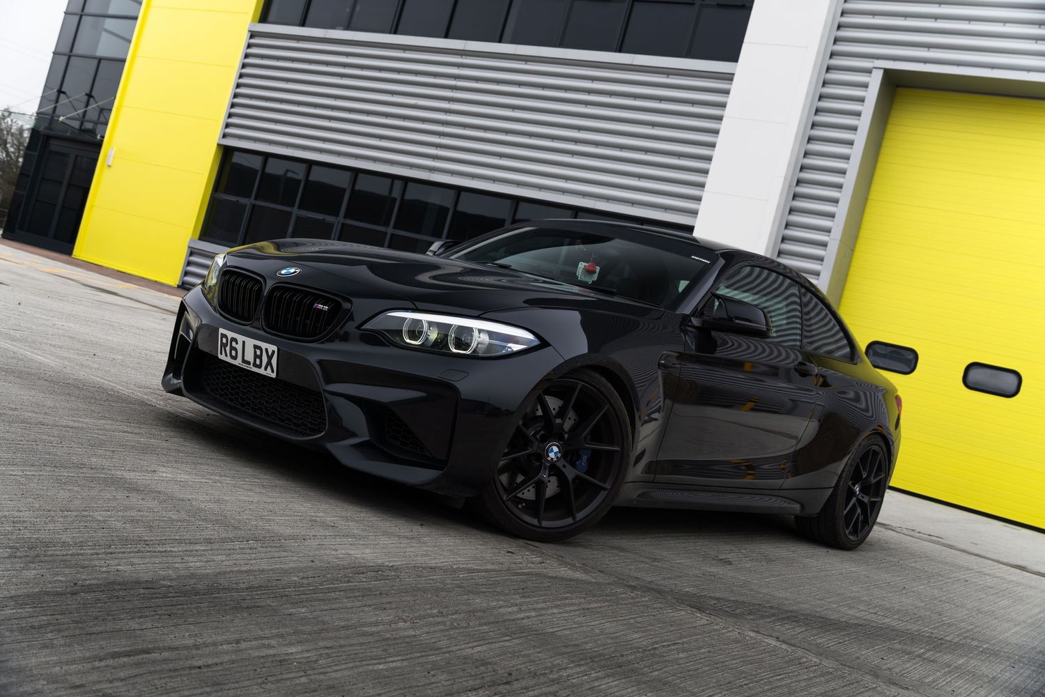 Black BMW M2 coupe parked in front of yellow garage doors at an industrial unit.