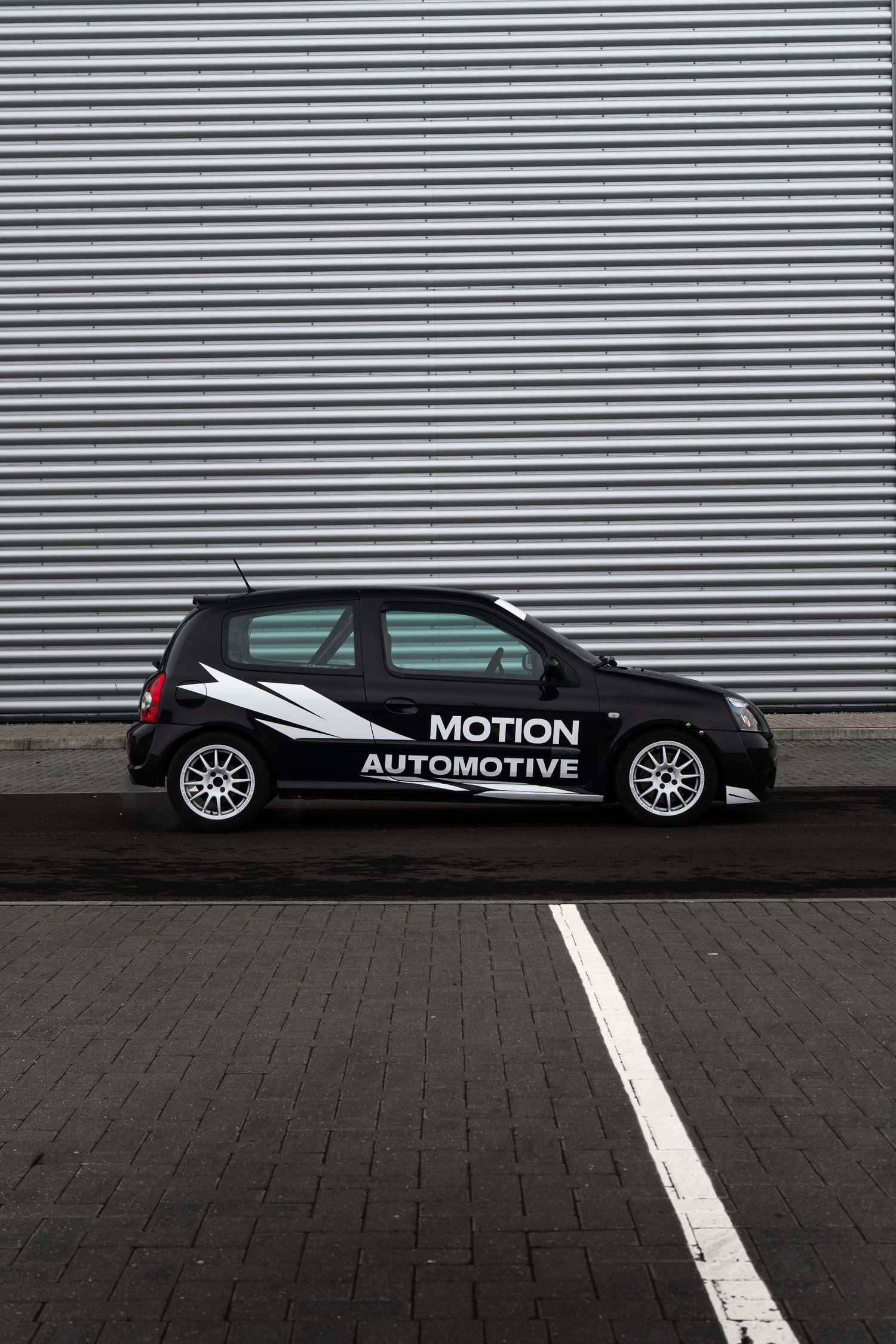 Black Motion Automotive branded Renault Clio sport car parked against corrugated metal wall.