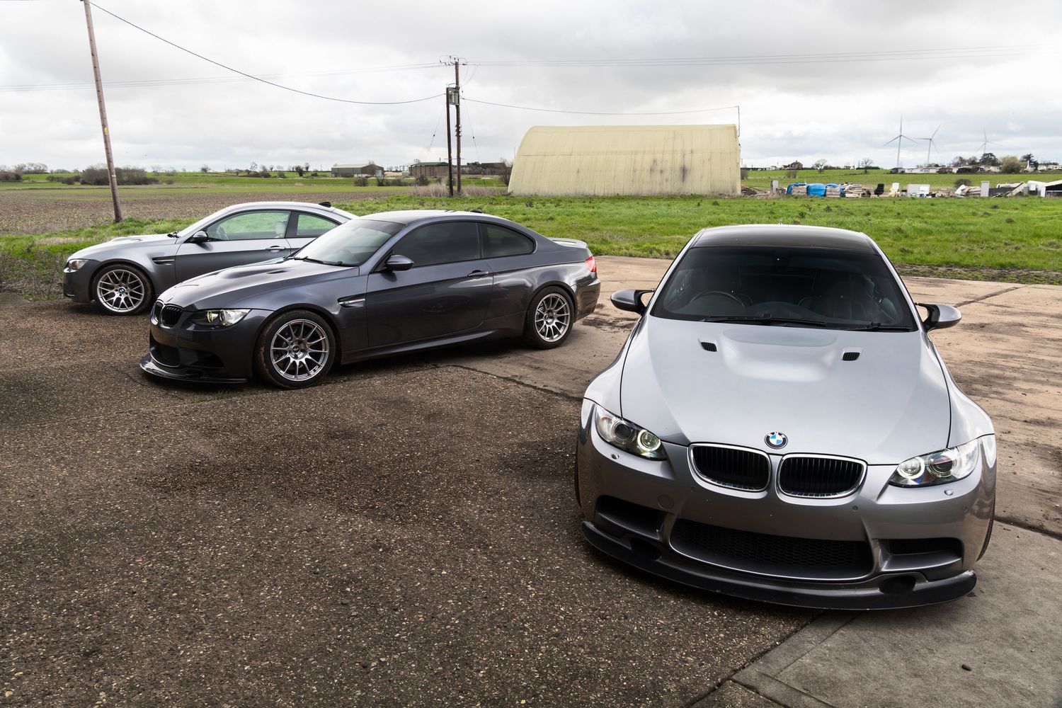 Three BMW M3 cars parked on gravel lot near green field, silver M3 prominently in foreground.