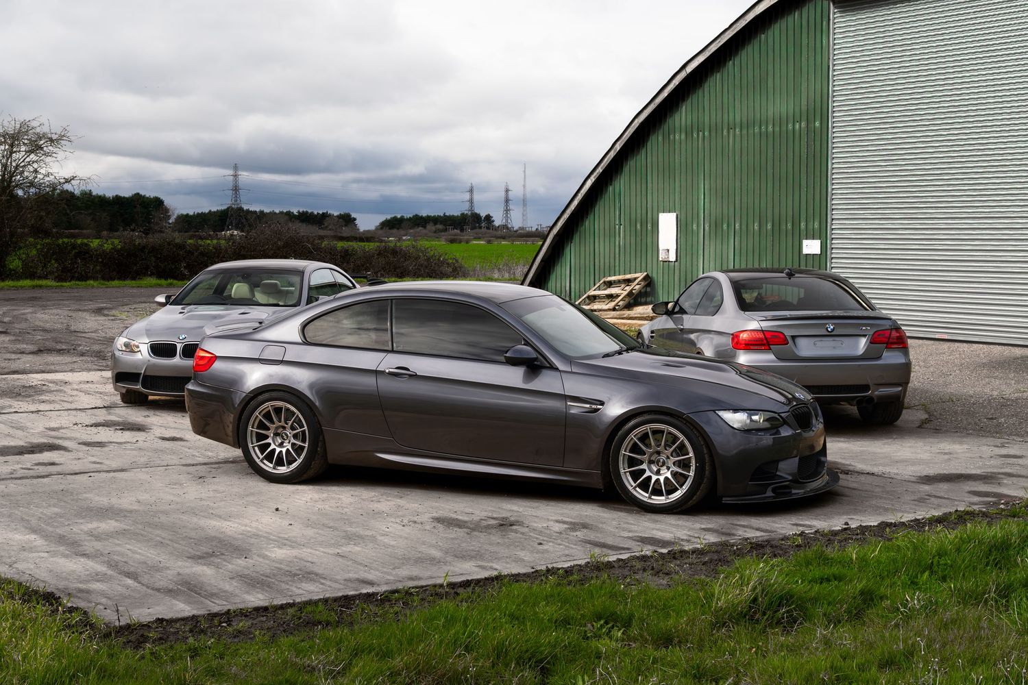 Three gray BMW M3 coupes arranged outside a large green arched metal warehouse building.