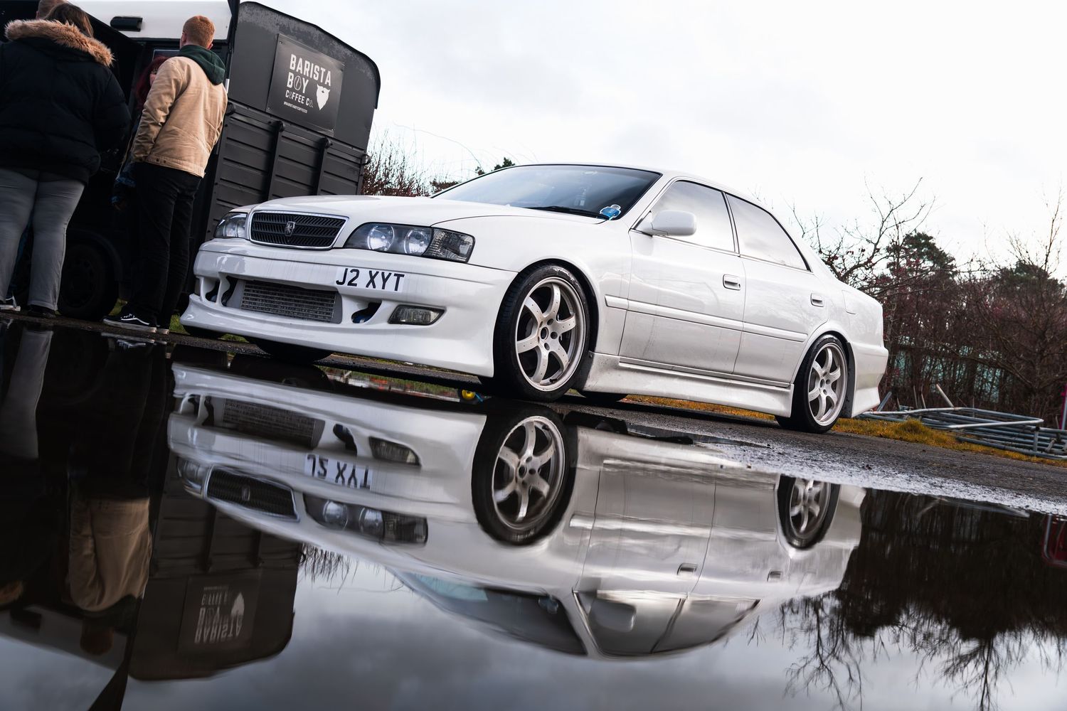 White Toyota Chaser JZX100 reflected in puddle at a car show event.