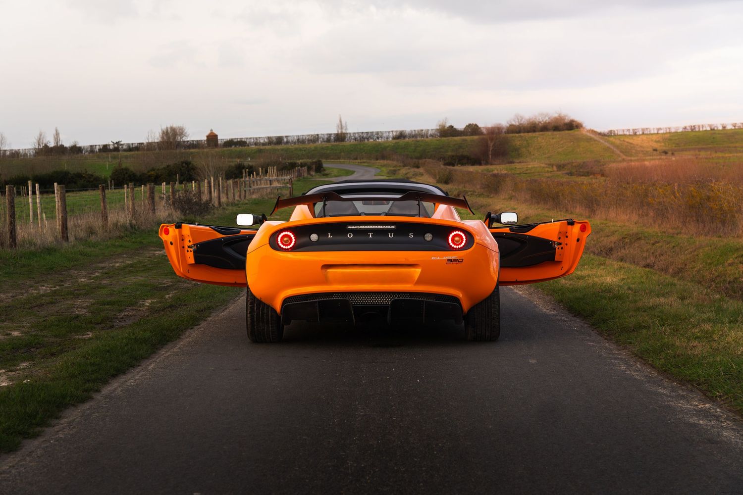 Rear view of orange Lotus Elise sports car on rural road with open doors.