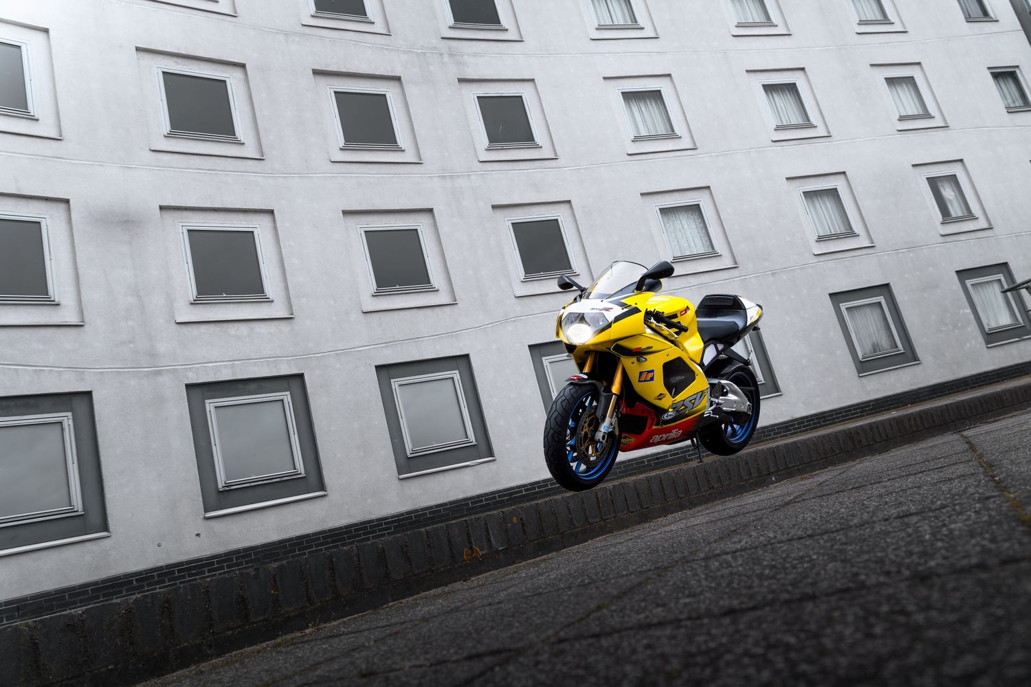 Yellow Aprilia RSV sportbike leaning on a road beside a large white multi-story building.