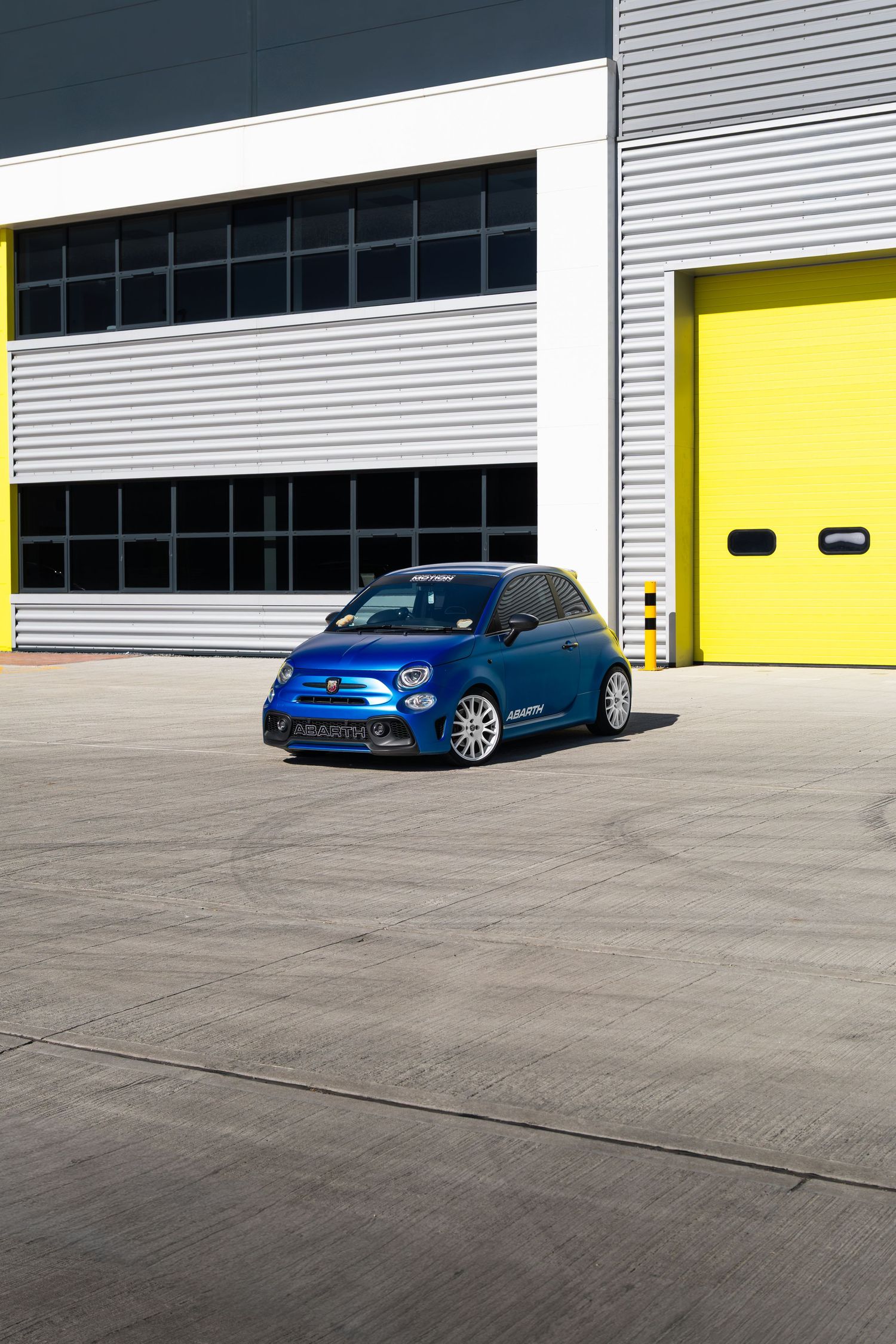 Blue Abarth 500 performance hatchback parked on tarmac in front of yellow garage doors.