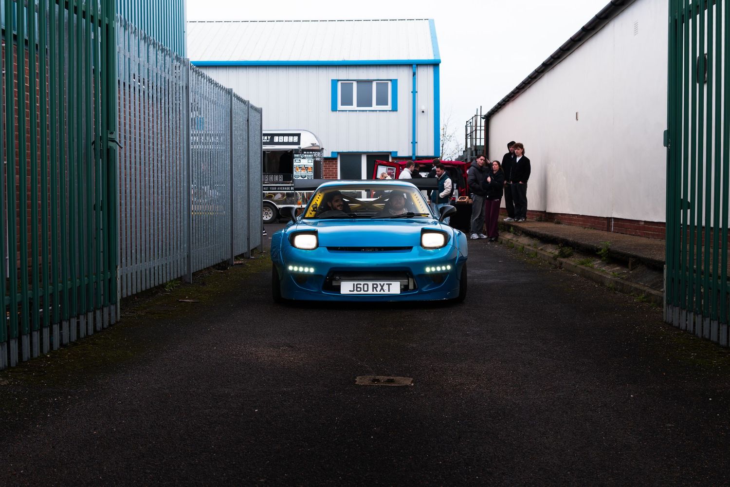 A modified blue sports car with headlights on drives through a narrow industrial gate entrance.