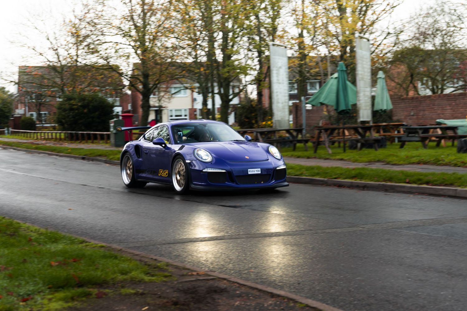 A blue Porsche 911 GT3 driving on a wet road past a pub car park.