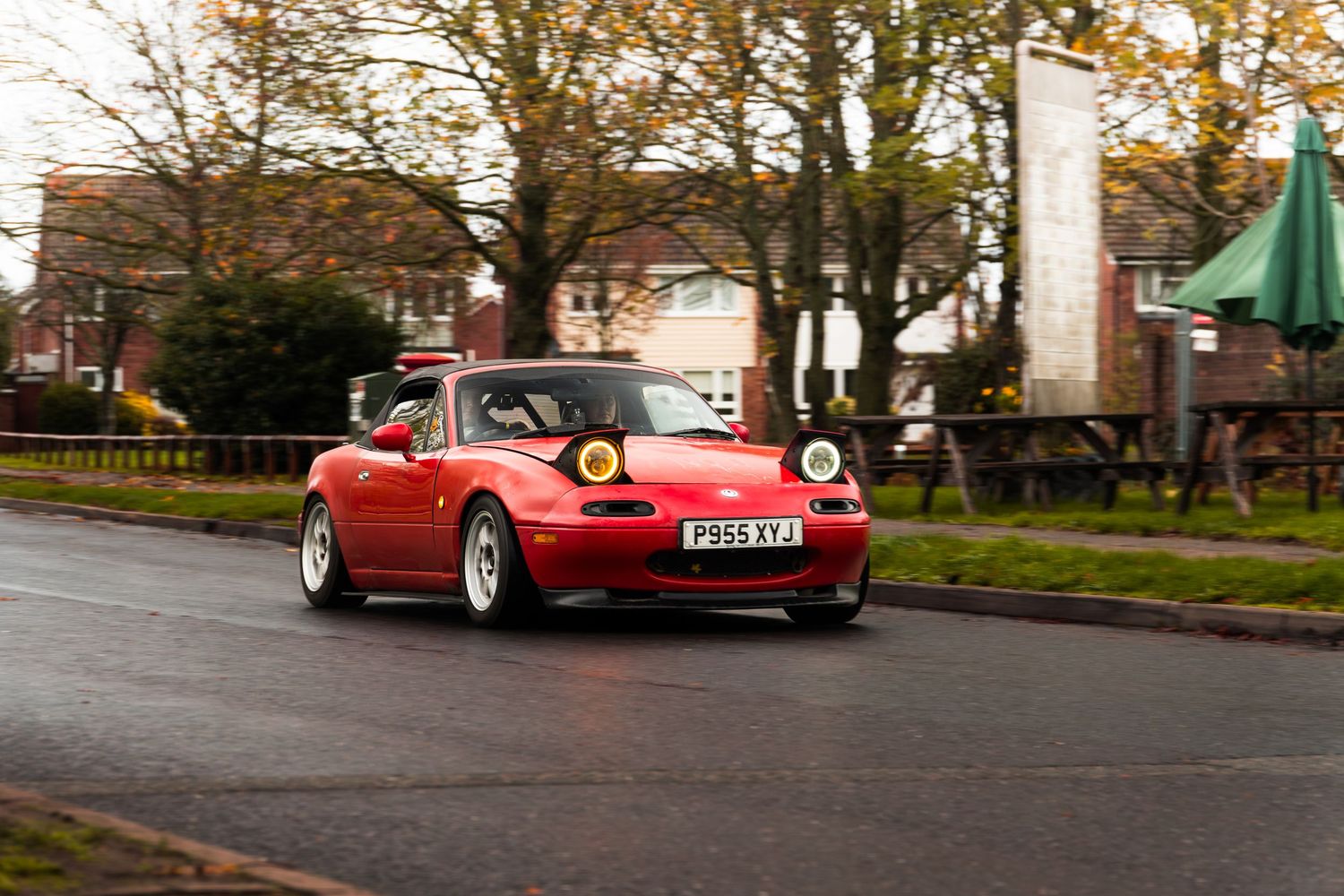 Red classic Mazda MX-5 NA with white wheels driving on wet UK road in autumn.