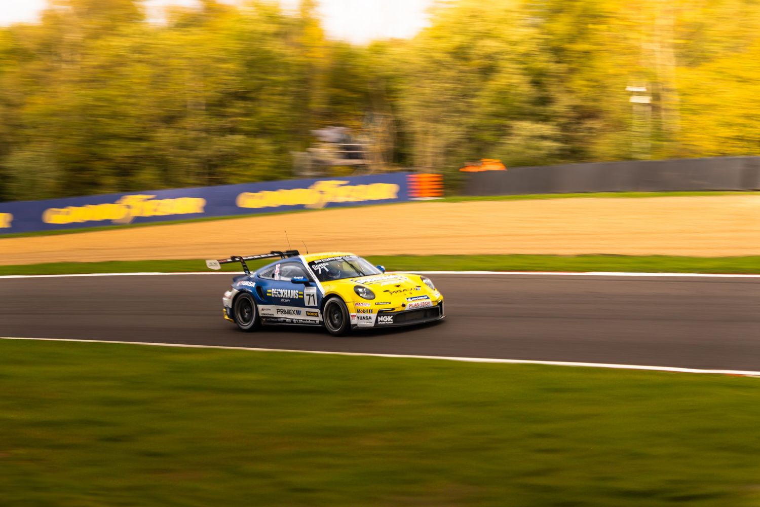 Blue and yellow Porsche GT3 Cup race car speeding on circuit with golden evening light background.