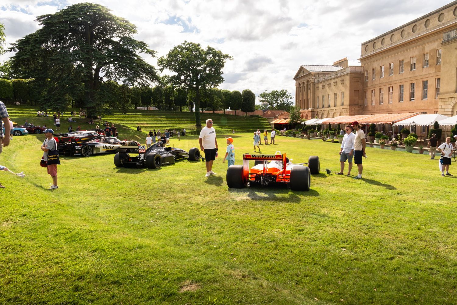 Formula 1 cars displayed on green lawn at historic estate with visitors walking around during outdoor motorsport event.