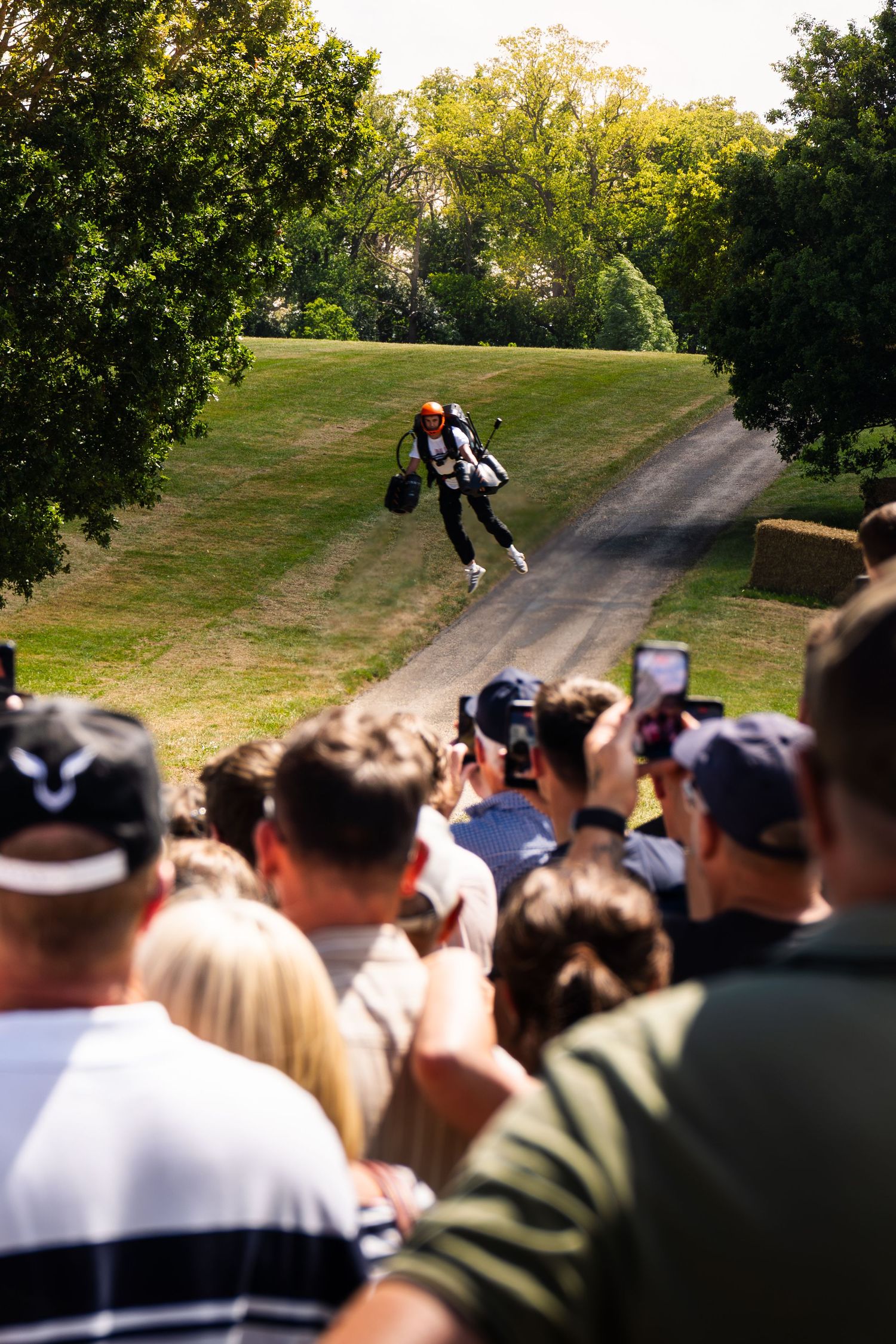 Jetpack pilot flies over a crowd of spectators filming with phones at an outdoor event on a sunny day.