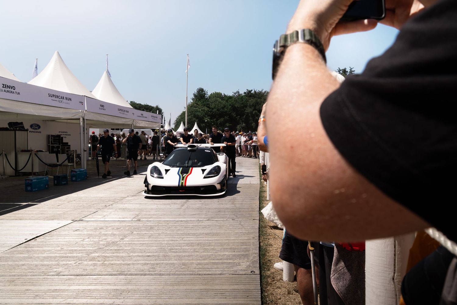 White racing supercar on display at an outdoor motorsport event with spectators photographing it on a sunny day.