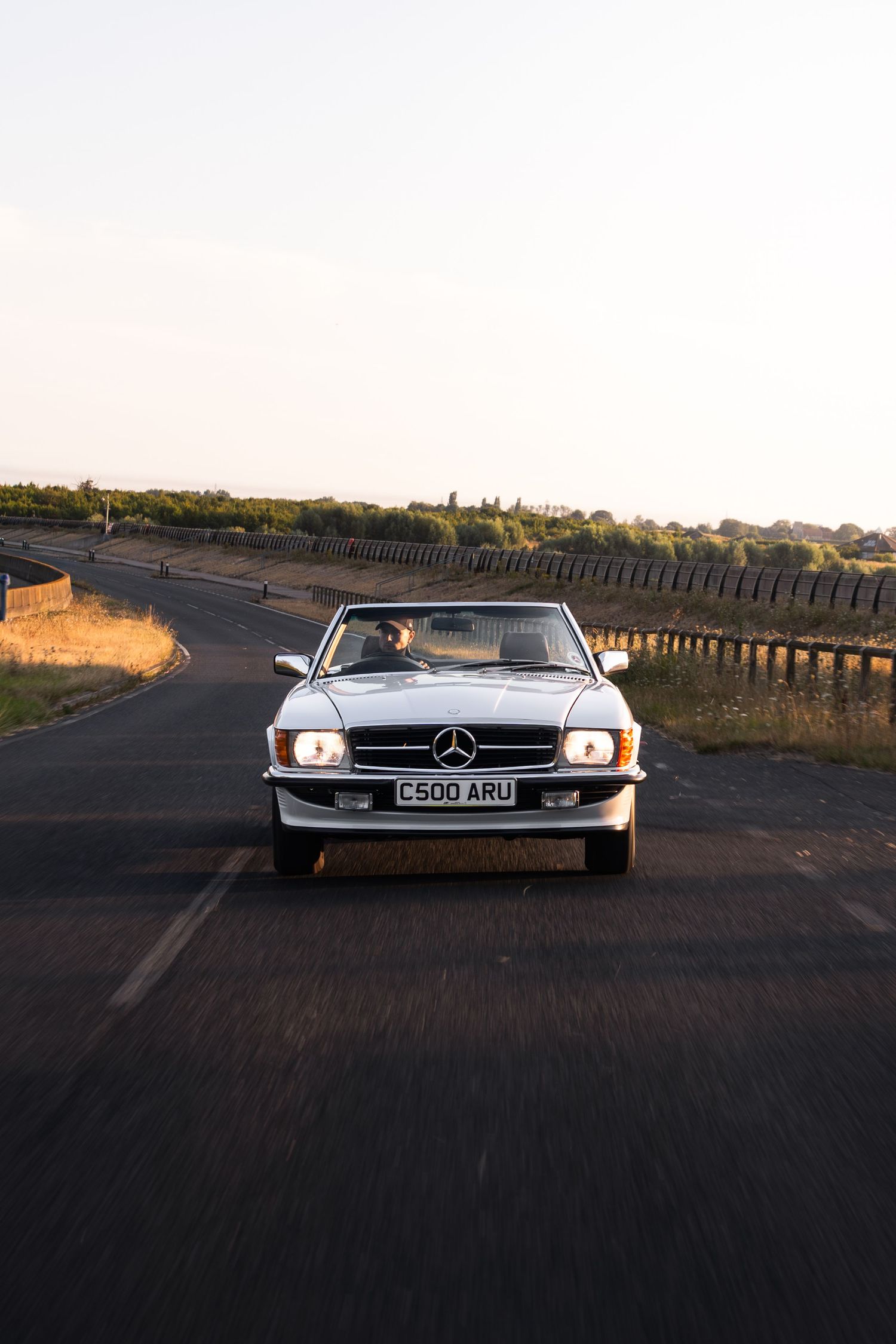 Classic white Mercedes-Benz convertible driving on a rural road at golden hour, UK license plate C500 ARU.
