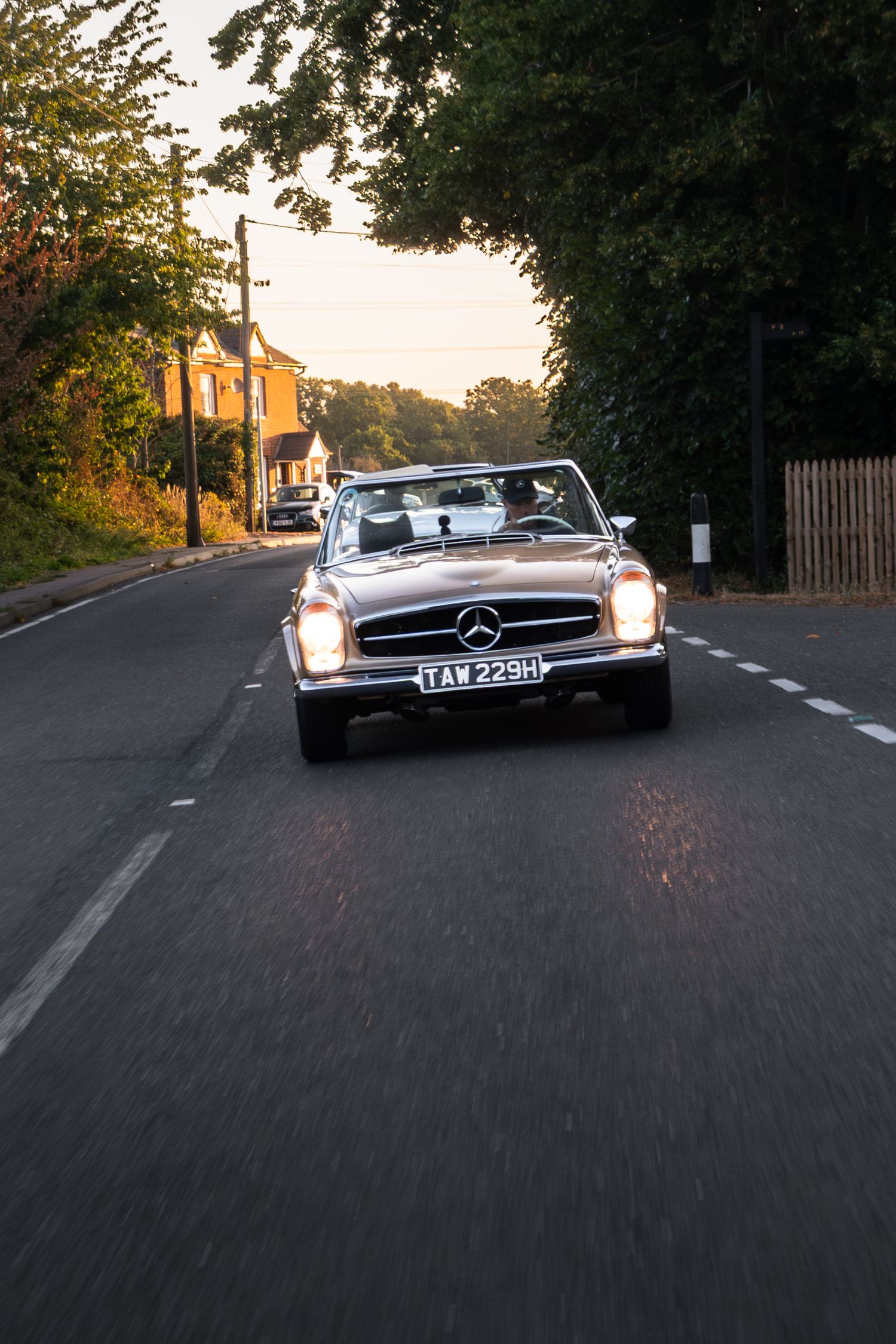 Classic Mercedes-Benz convertible driving on a tree-lined road at sunset with headlights on.