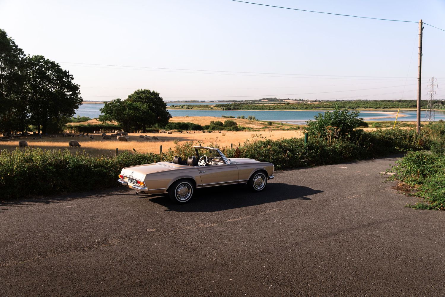 Vintage white convertible car parked on a rural road with scenic water views and golden fields in the background.