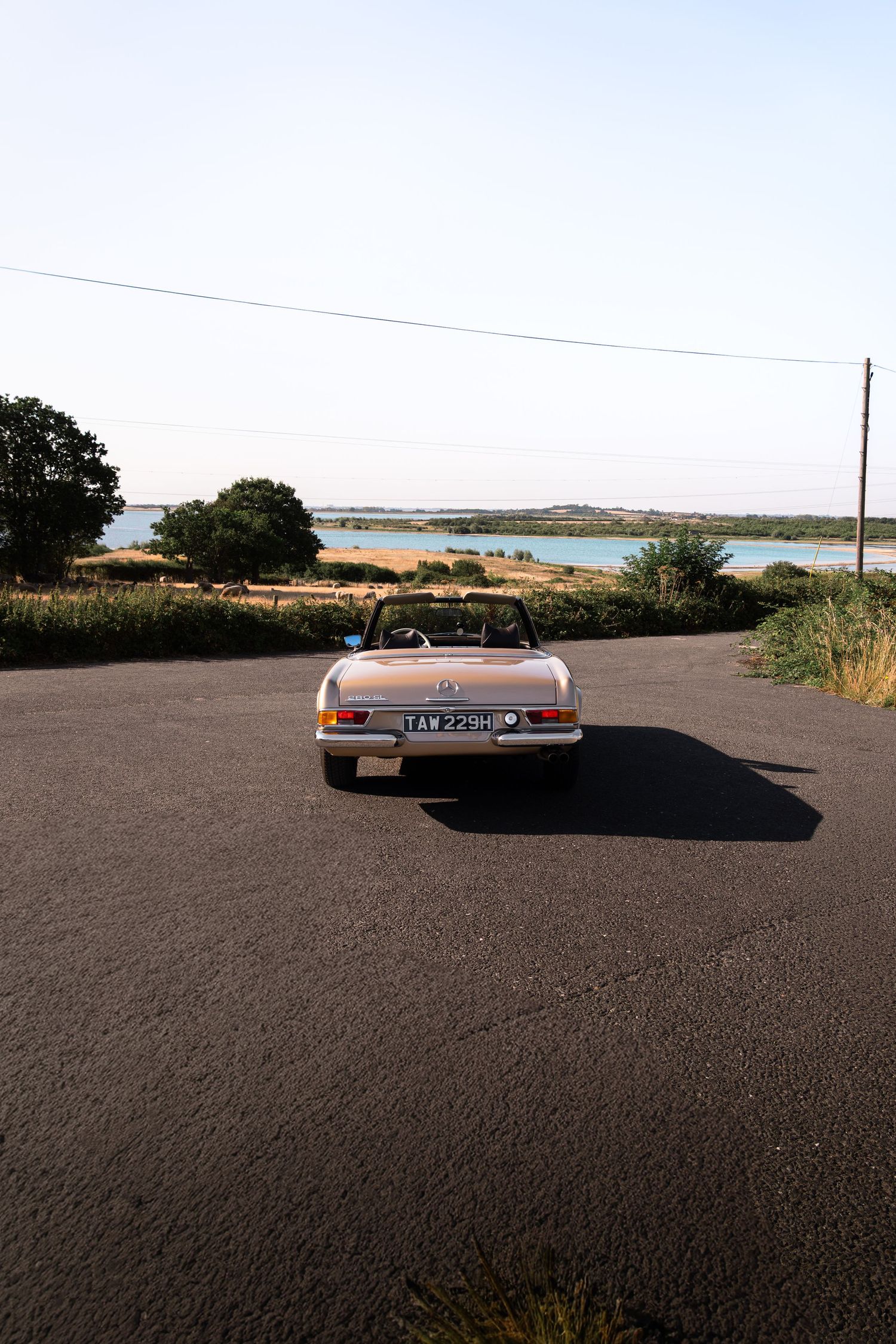 Vintage convertible car parked on a rural road with scenic water views and trees under a clear sky.