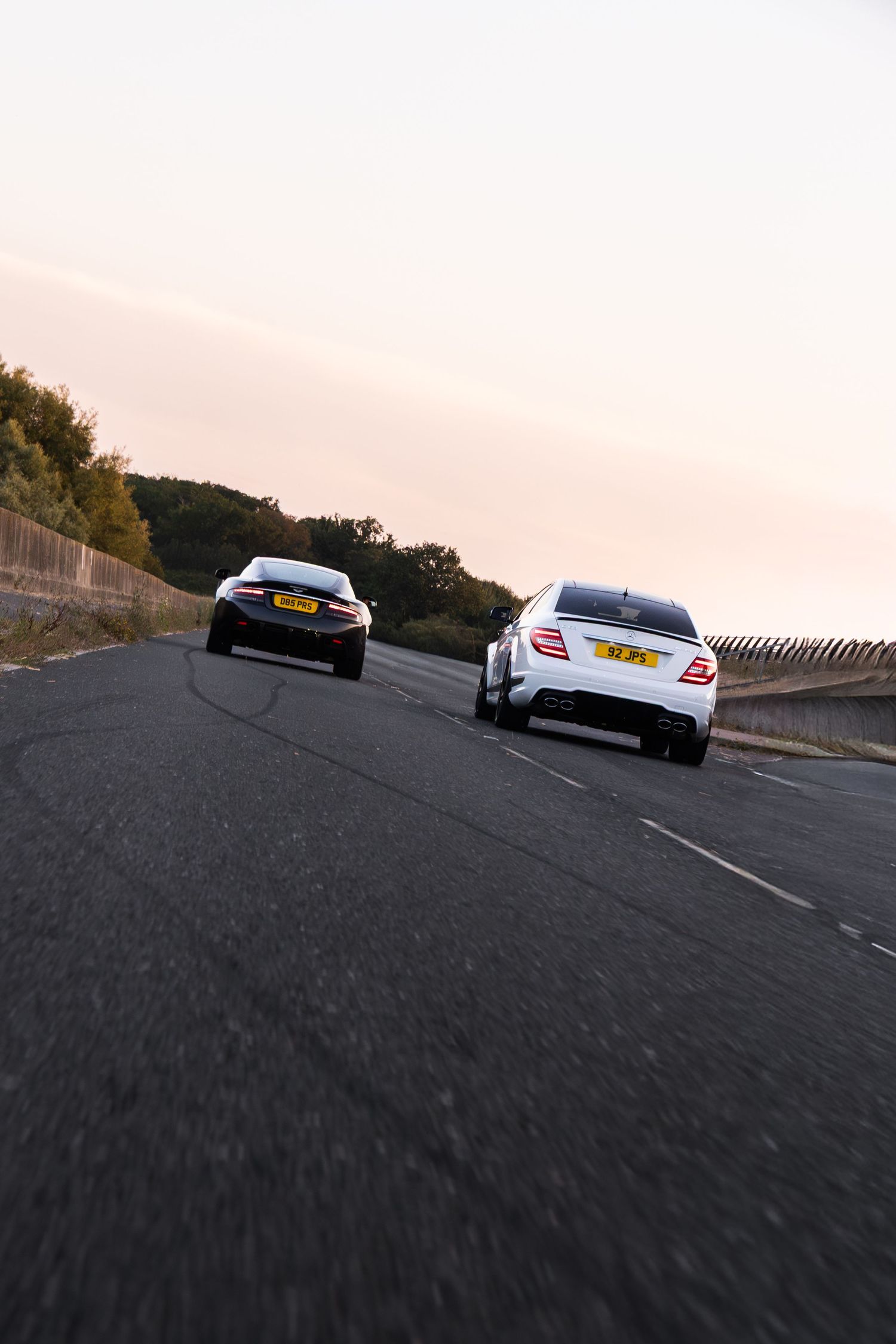 Two cars racing on a road at dusk, white Mercedes and black sports car with UK license plates on a curved highway.